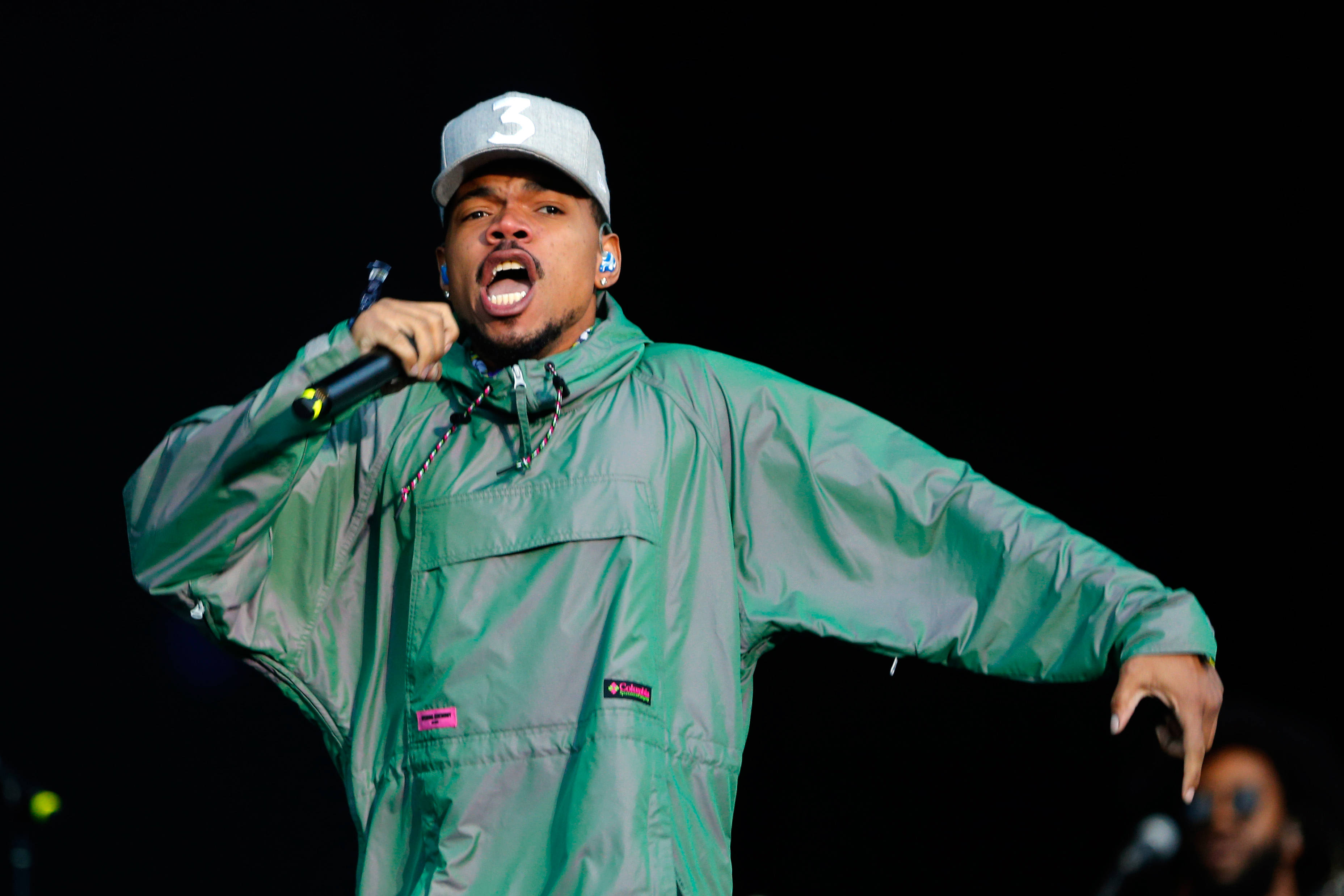 chance 桌布 壁紙 — 一位身穿綠色外套和白色帽子的男性饒舌歌手，在黑暗的舞台背景下熱情地對著麥克風表演。