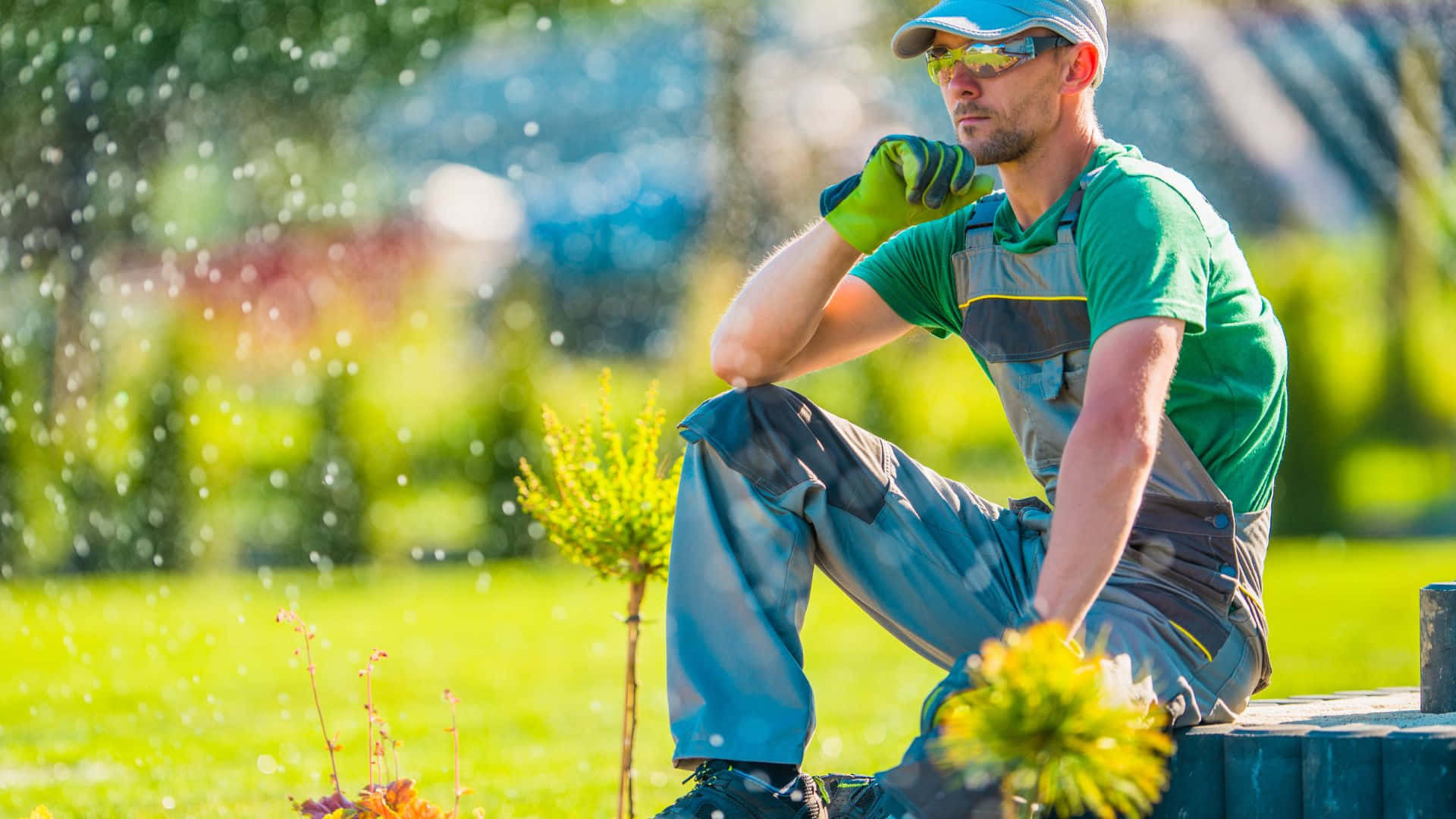 Garden Design 桌布 壁紙 — 一位身穿綠色工作服和手套的園丁坐在木長椅上，陽光普照的花園中水珠在他周圍噴灑。