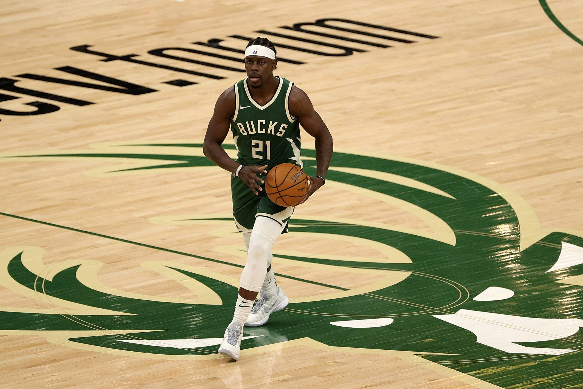 Jrue Holiday 桌布 壁紙 — 一位身穿綠色球衣的密爾瓦基公鹿籃球運動員在帶有球隊標誌的拋光木地板上運球。