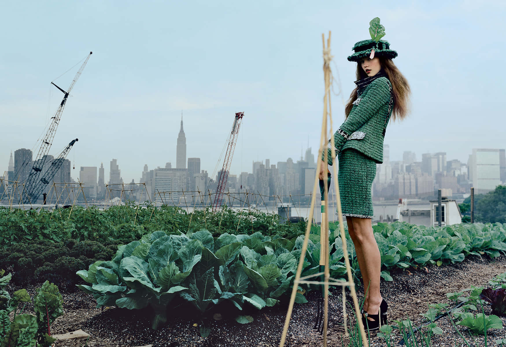 Green Fashion 桌布 壁紙 — 一位身穿綠色粗花呢套裝、戴著樹葉帽子的女士站在屋頂花園中，身後是朦朧的紐約天際線和建築起重機。