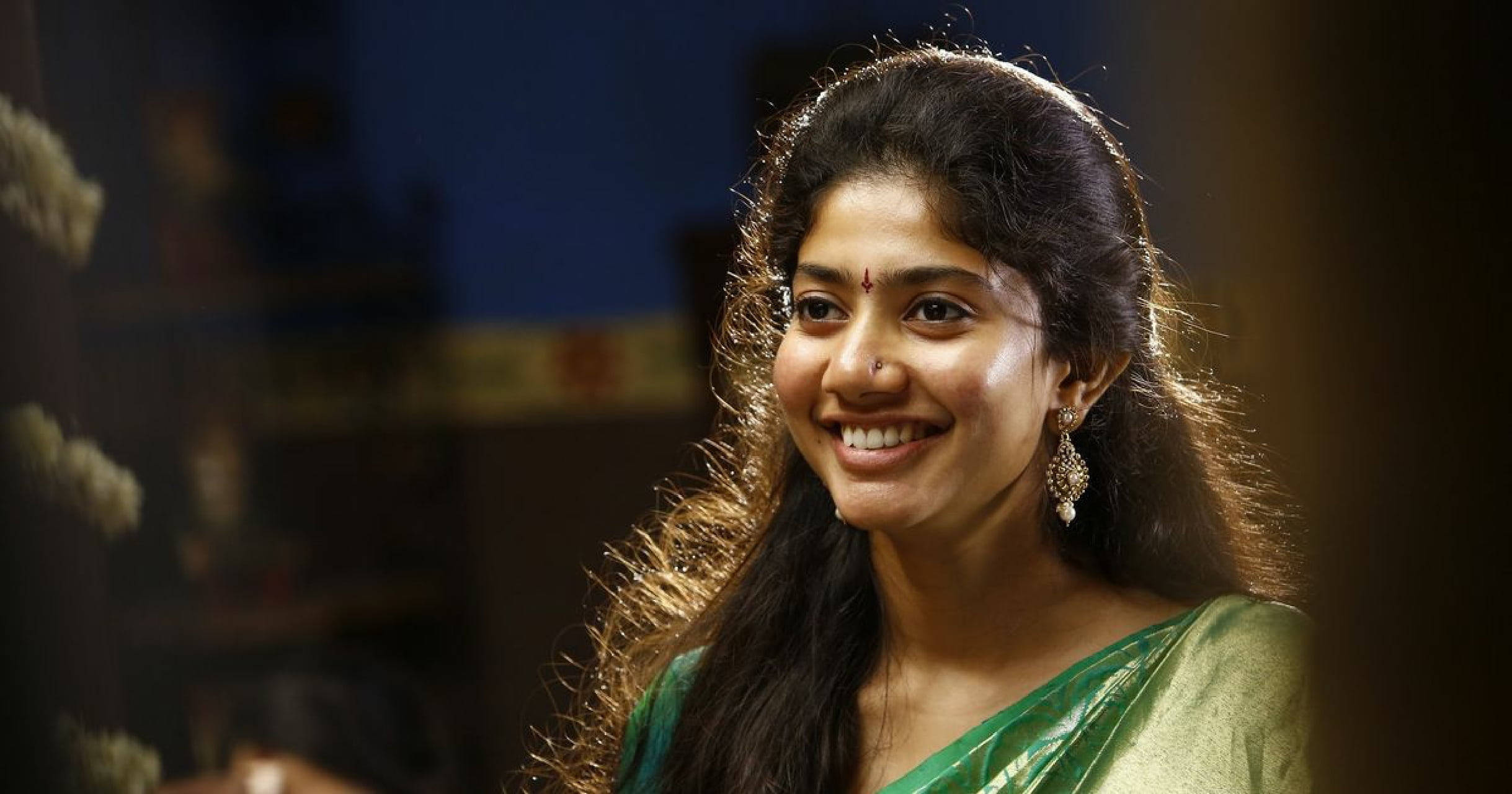 Sai Pallavi 桌布 壁紙 — 一位身穿綠色紗麗的容光煥發的女性，戴著傳統的眉心貼和耳環，在柔和模糊的室內背景下溫暖地微笑。