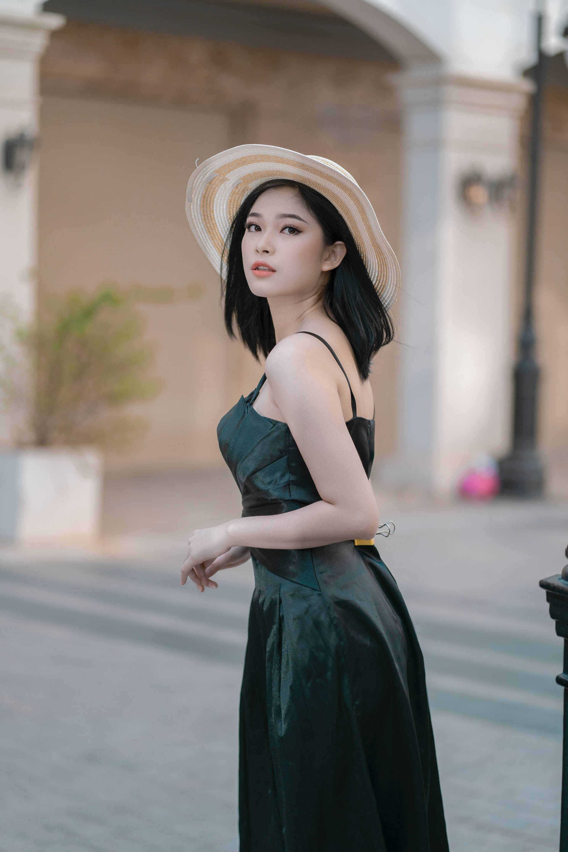 lovely girl 桌布 壁紙 — 一位身穿綠色緞面連衣裙和草帽的女性，在陽光普照的城市街道上，以柔和的建築背景優雅站立。