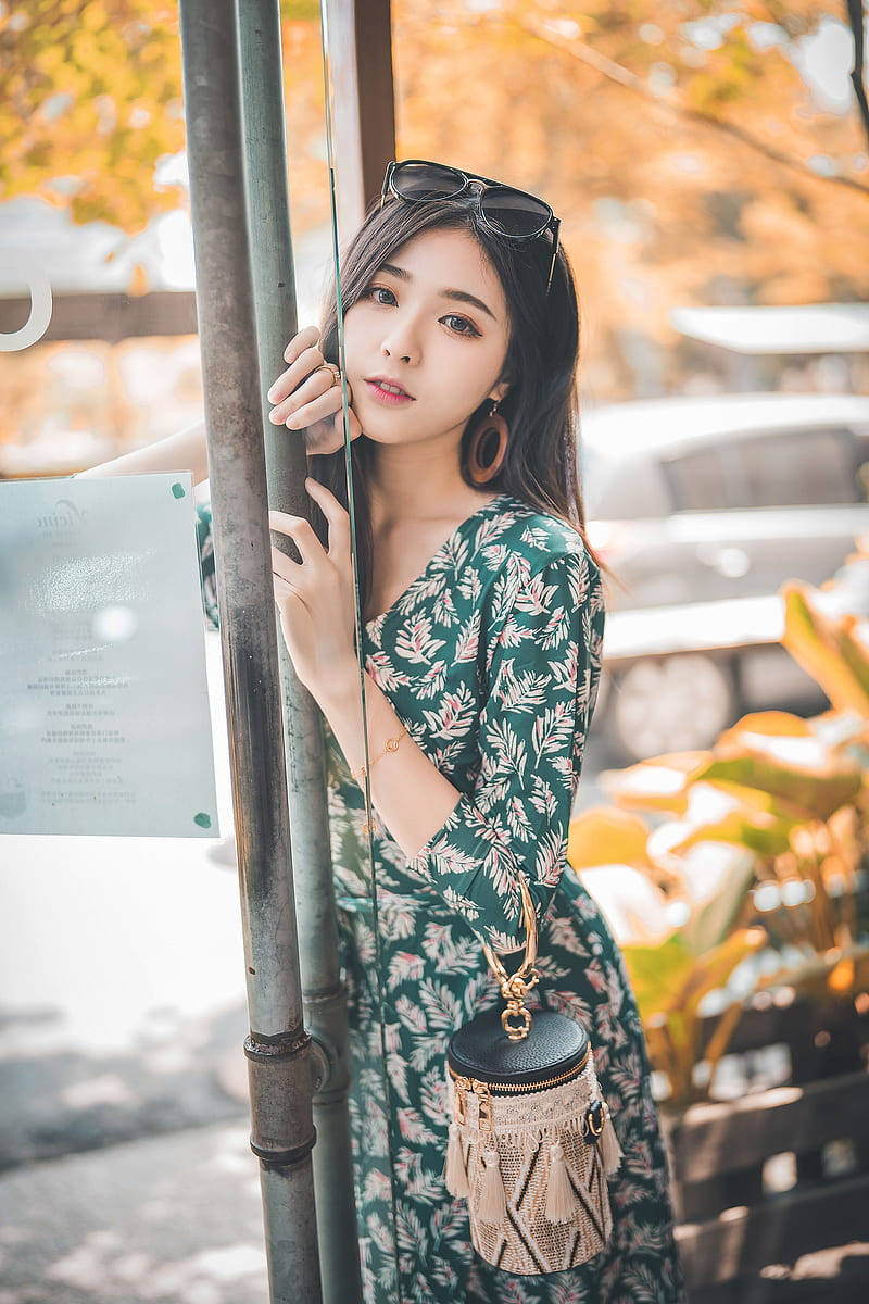美女 桌布 壁紙 — 一位身穿綠色花卉連衣裙的年輕女性在戶外靠著柱子，背景有秋葉和柔和陽光。