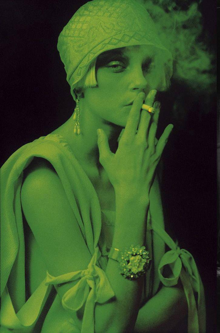 jessica stam 桌布 壁紙 — 一位身穿綠色調 flapper 連衣裙和鐘形帽的女性在深色背景下吸煙。