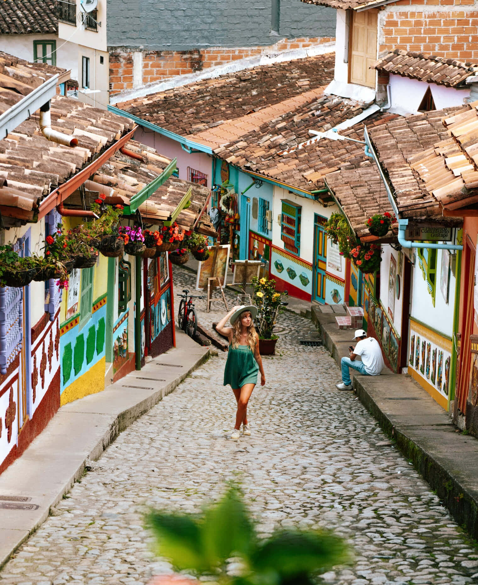 Colombia 桌布 壁紙 — 一位身穿綠色連衣裙的女子走在一條充滿活力的鵝卵石街道上，街道兩旁是色彩繽紛的殖民時期建築和懸掛的花籃。