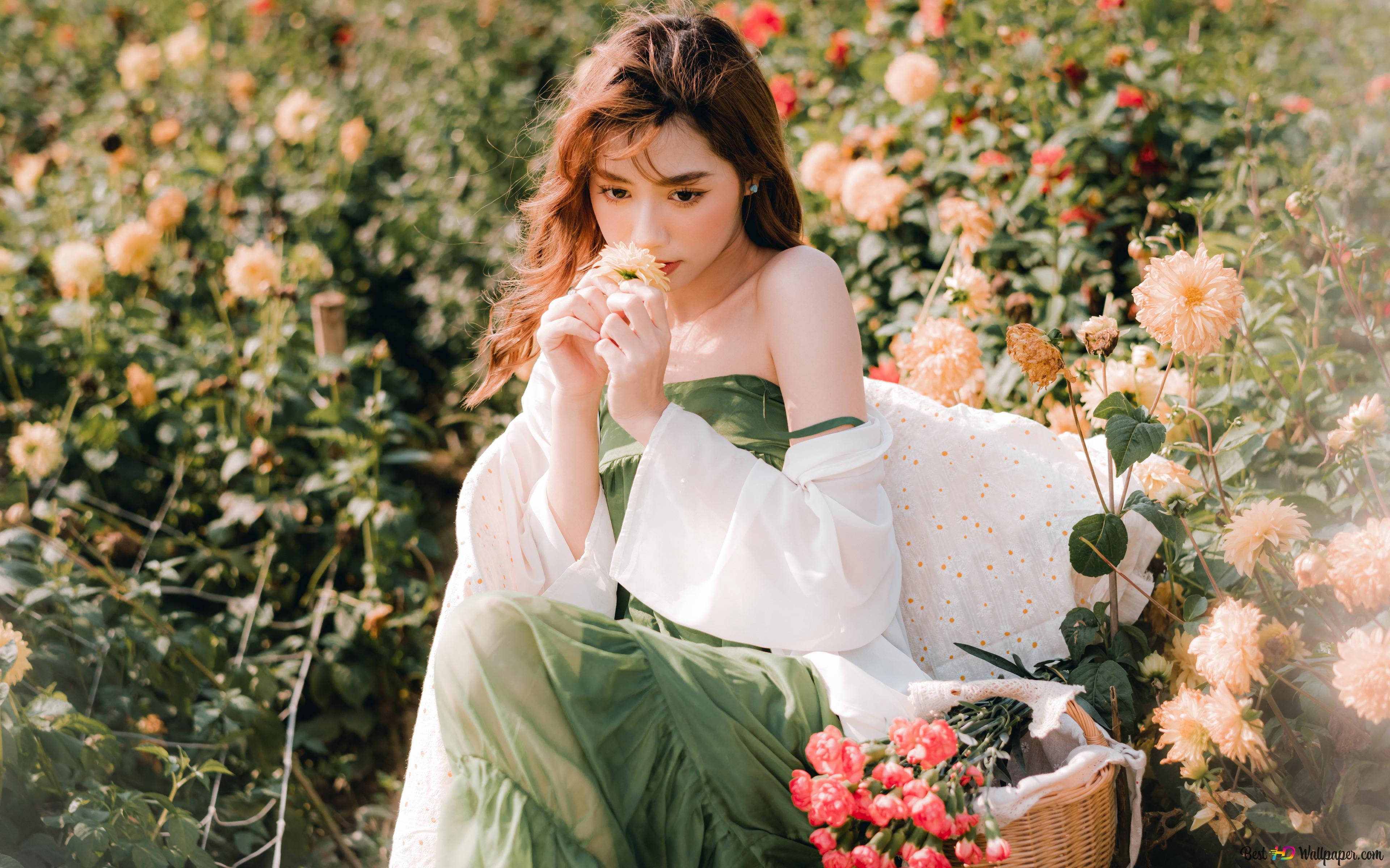 美女 桌布 壁紙 — 一位身穿綠色連衣裙的年輕女子坐在一片桃色和紅色花朵的田野中，將一朵小花湊到鼻尖。