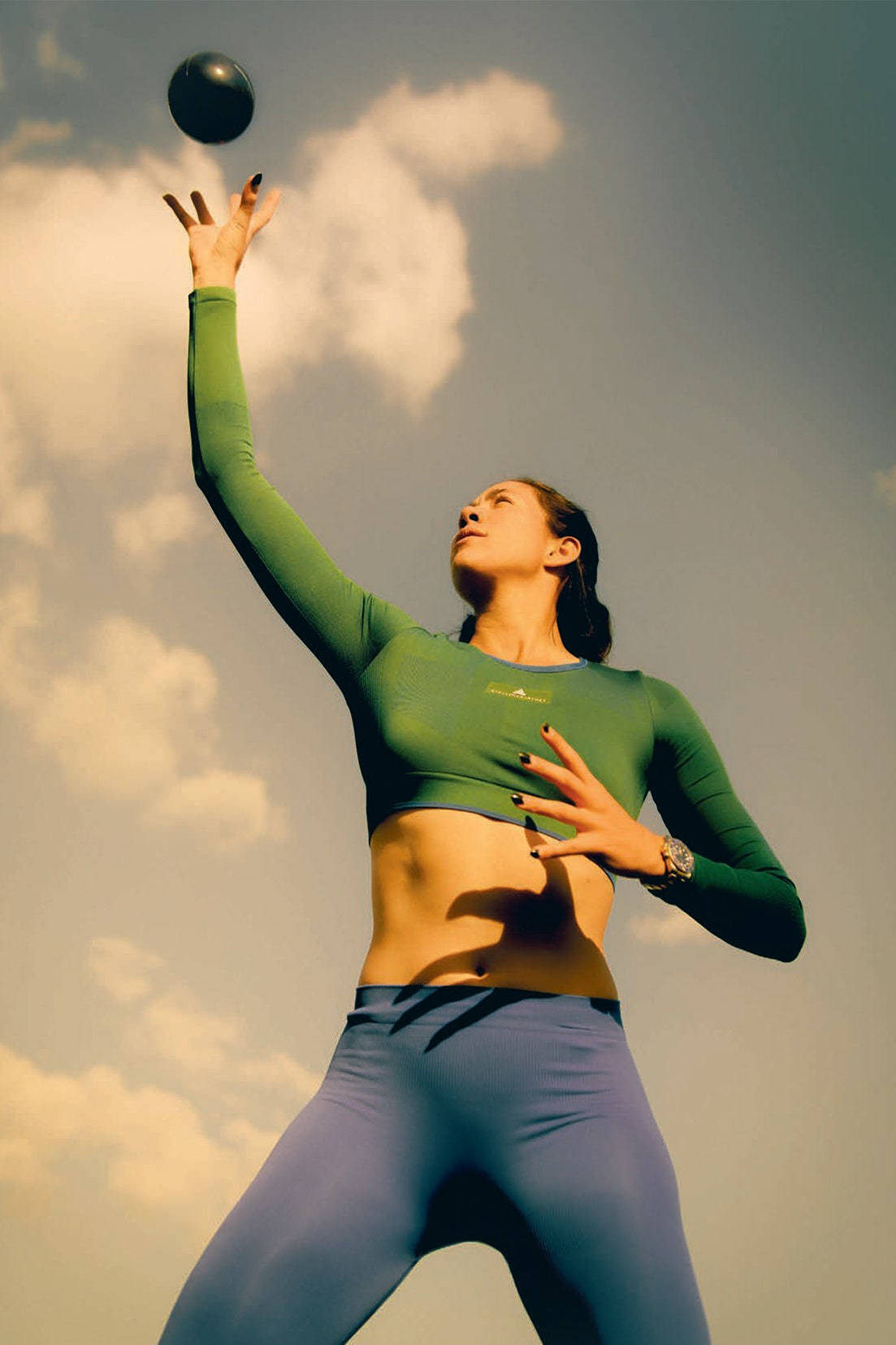 Athlete 桌布 壁紙 — 一位身穿綠色運動服的女士在溫暖的金色光線下，向多雲的天空投擲一個黑色球。