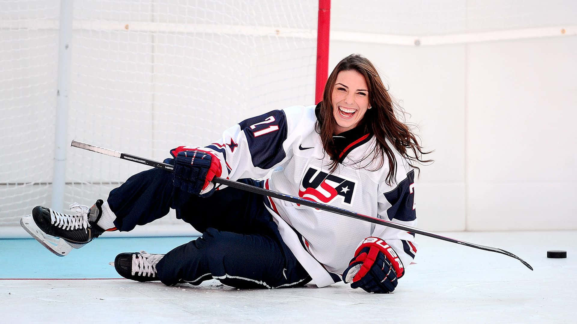 hockey 桌布 壁紙 — 一位身穿美國球衣的快樂女子冰球運動員，笑著用膝蓋在冰場上滑行，手中拿著球桿。