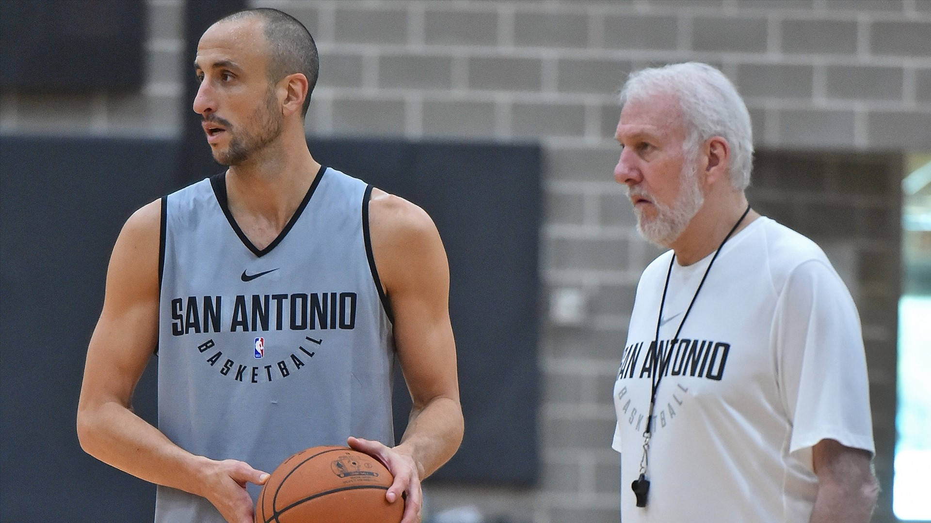 Manu 桌布 壁紙 — 一位身穿聖安東尼奧球衣的籃球運動員在磚牆體育館中拿著球與教練交談。