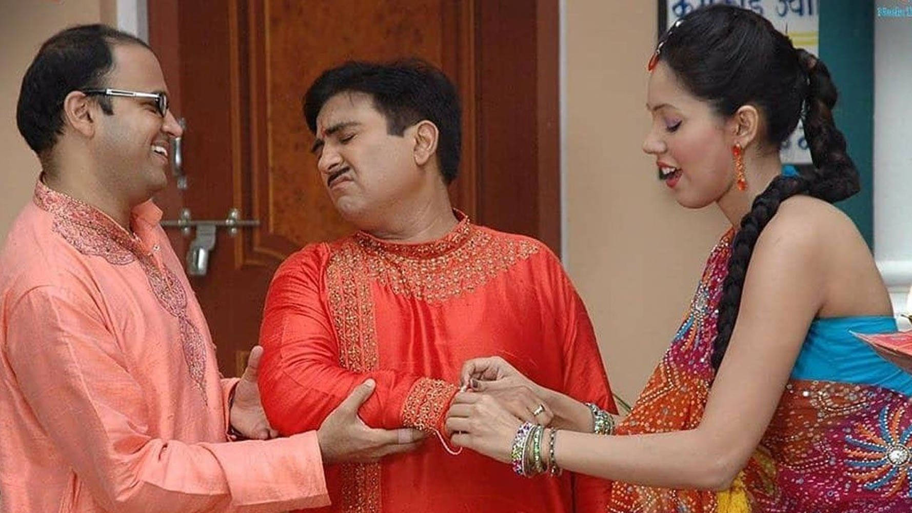 Celebrity 桌布 壁紙 — 一位身穿色彩繽紛刺繡紗麗的女子正在為一名男子的手腕塗上指甲花，另一名男子微笑觀看，場景在溫暖的室內燈光下。