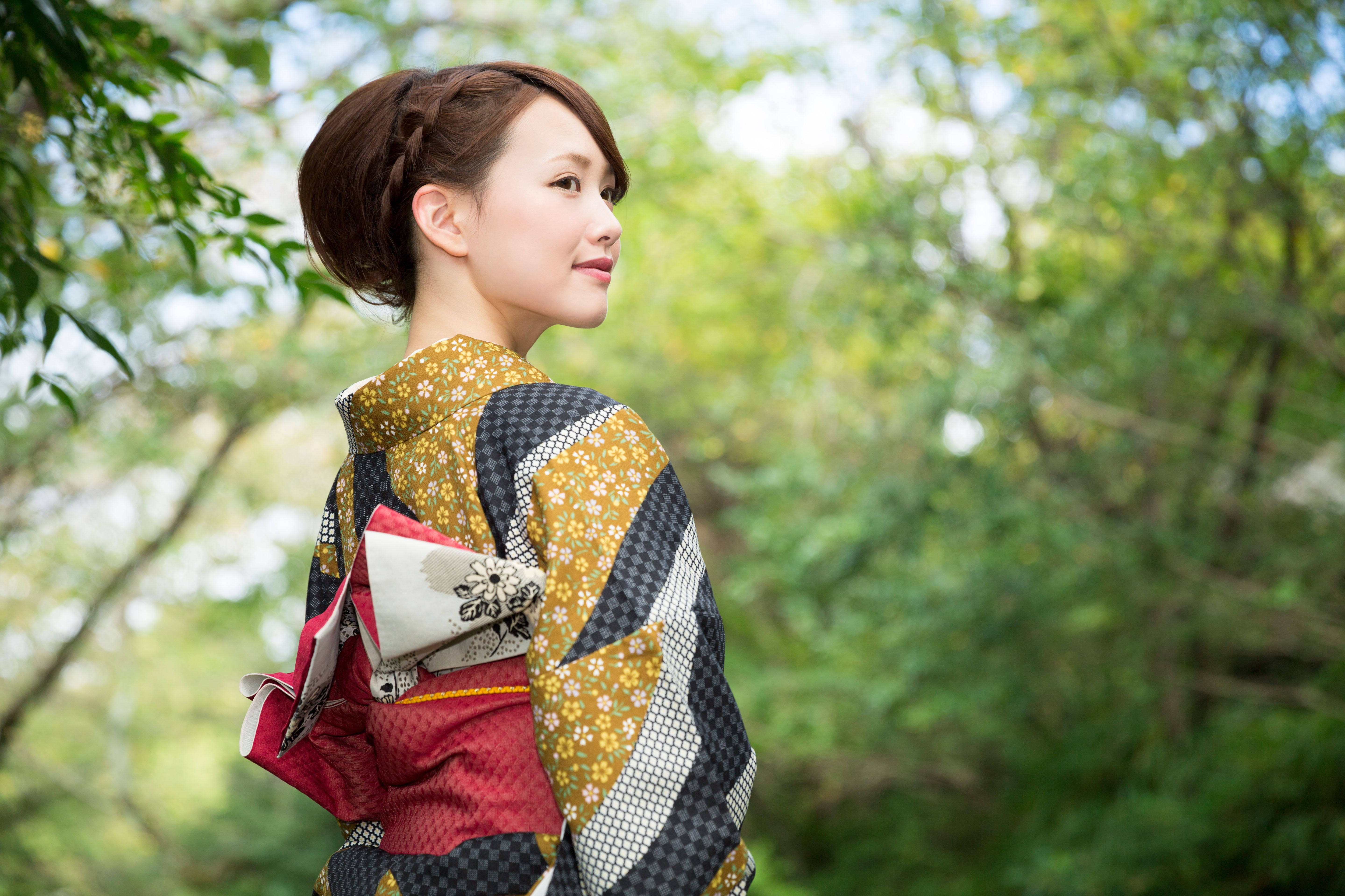 日本 桌布 壁紙 — 一位身穿色彩繽紛圖案和服的女人站在陽光普照的森林中，綠色樹葉在她身後柔和地模糊。