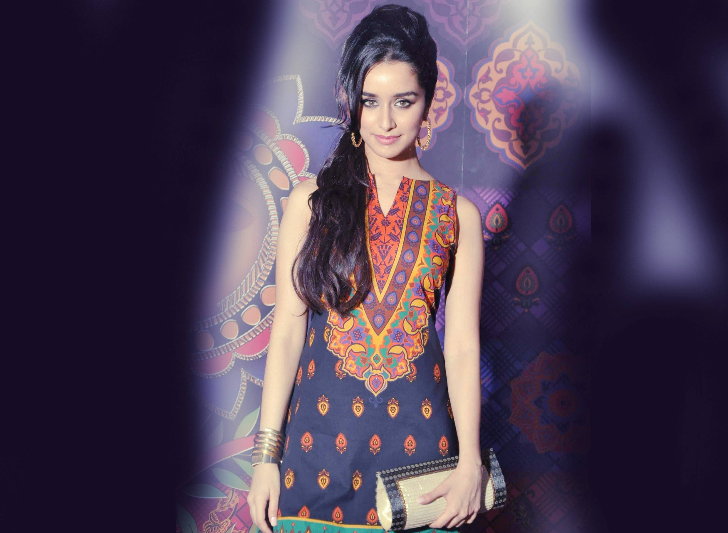 Shraddha Kapoor 桌布 壁紙 — 一位身穿色彩繽紛圖案連衣裙的女性自信地站在充滿活力的華麗背景前，帶有柔和聚光燈。