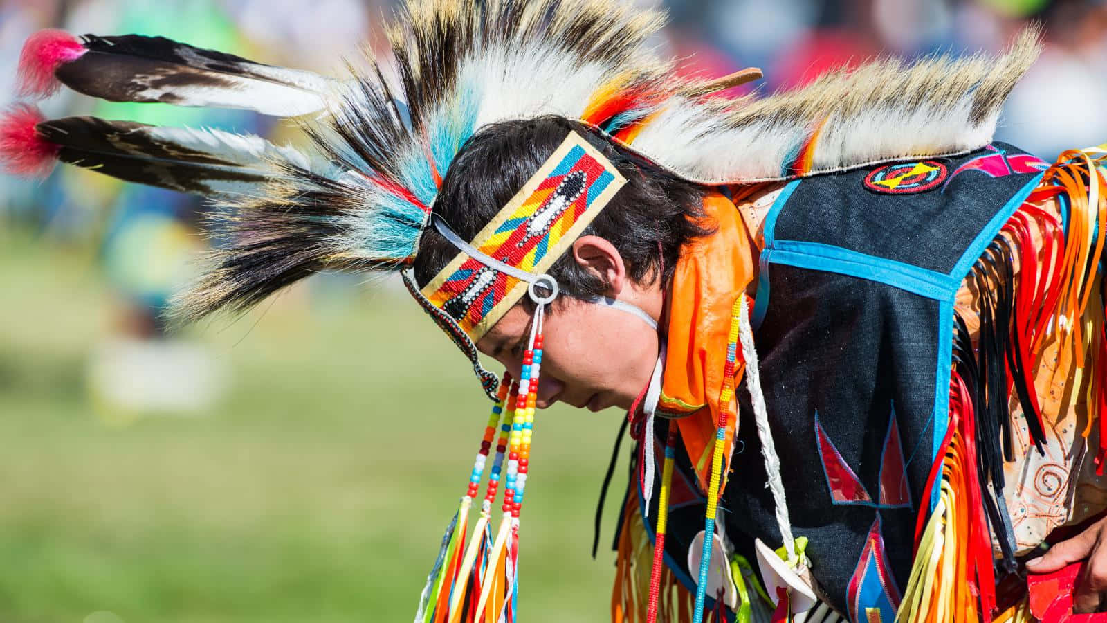 Native American 桌布 壁紙 — 一位身穿色彩鮮豔的傳統服飾、飾有羽毛和珠飾的人，在戶外低著頭。