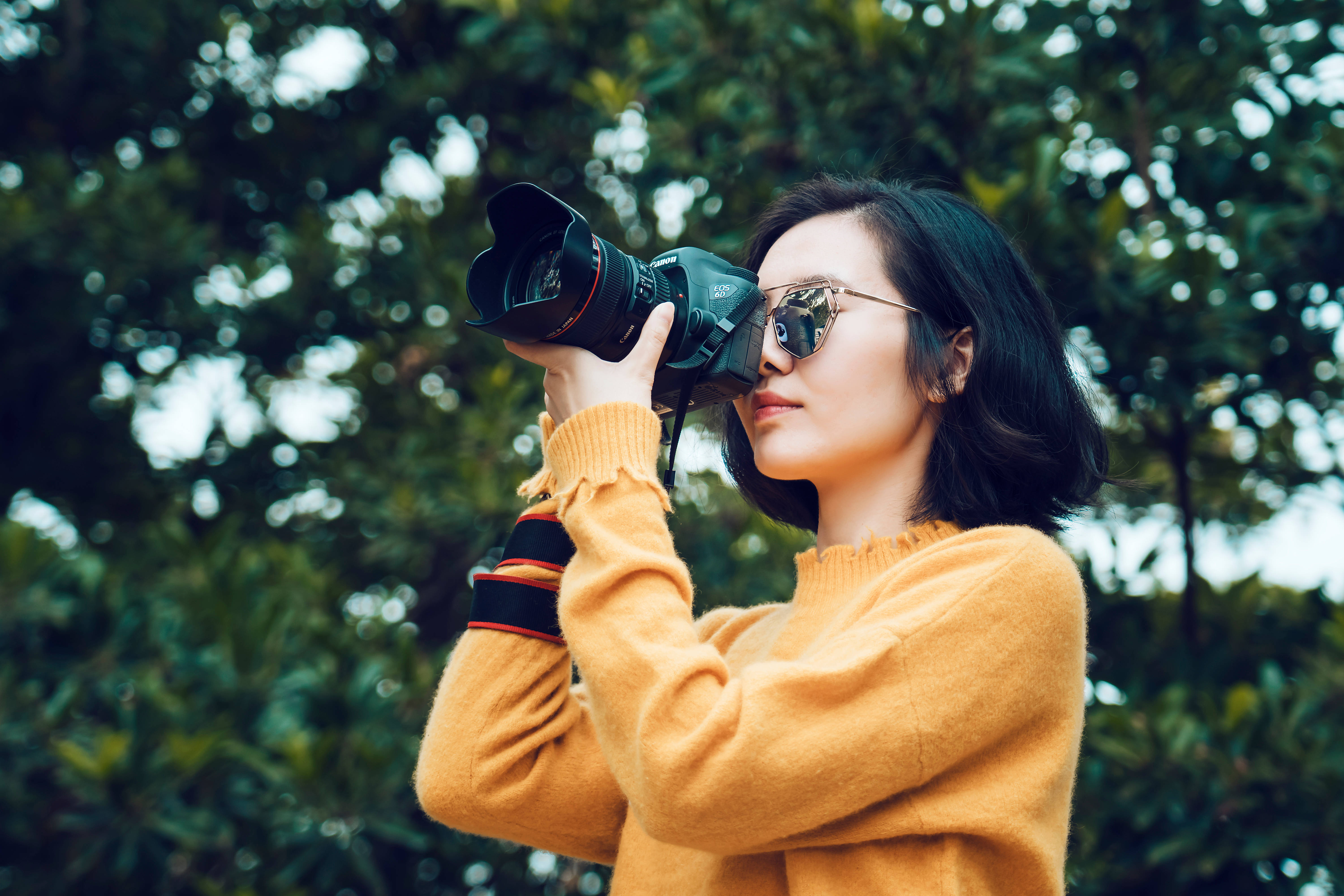 攝影 桌布 壁紙 — 一位身穿芥末黃毛衣和太陽眼鏡的女性，透過柔焦散景的森林背景瞄準單反相機。