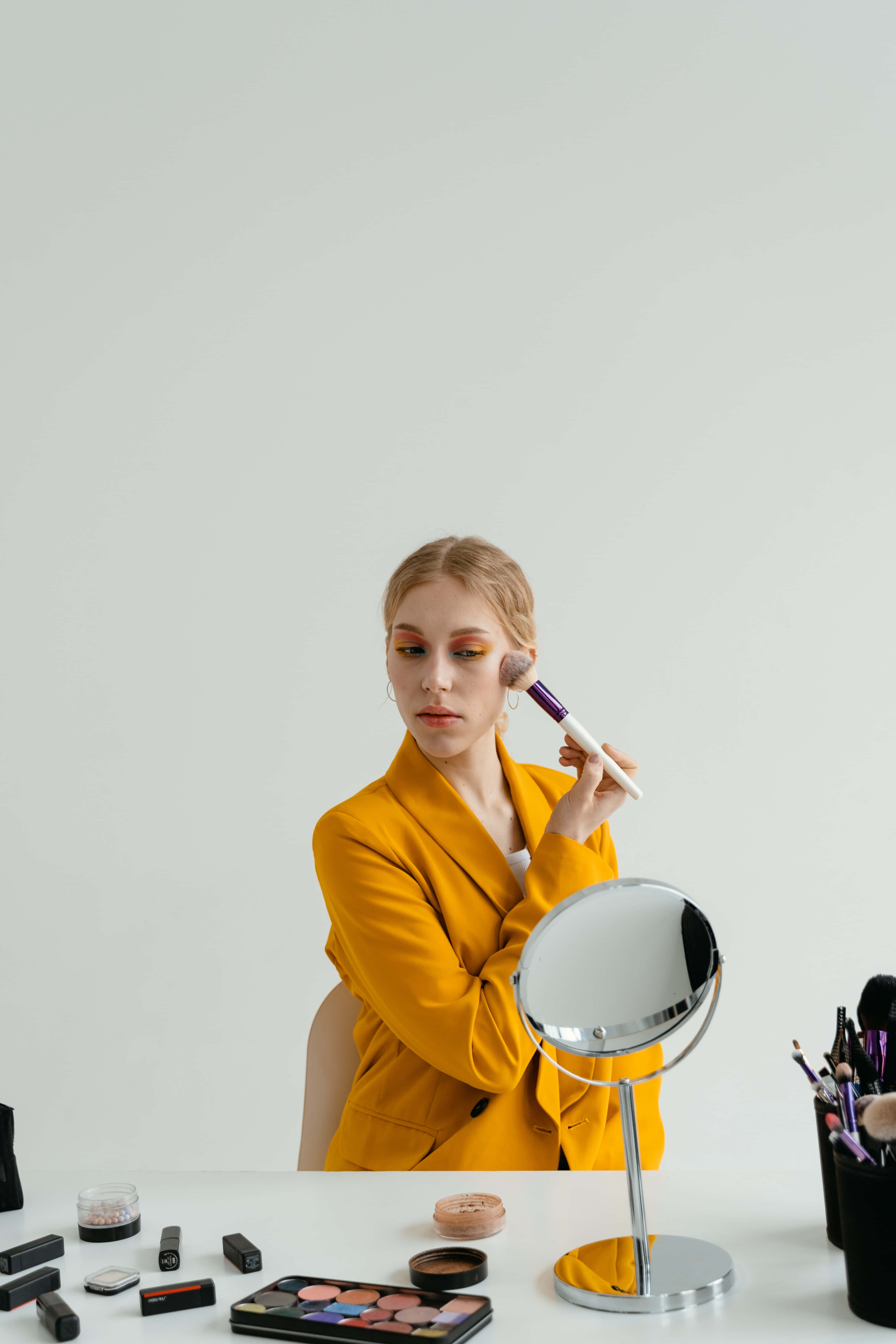 mirror 桌布 壁紙 — 一位身穿芥末黃色西裝外套的女性，坐在白色桌子旁，周圍是化妝品和圓鏡，用刷子化妝。