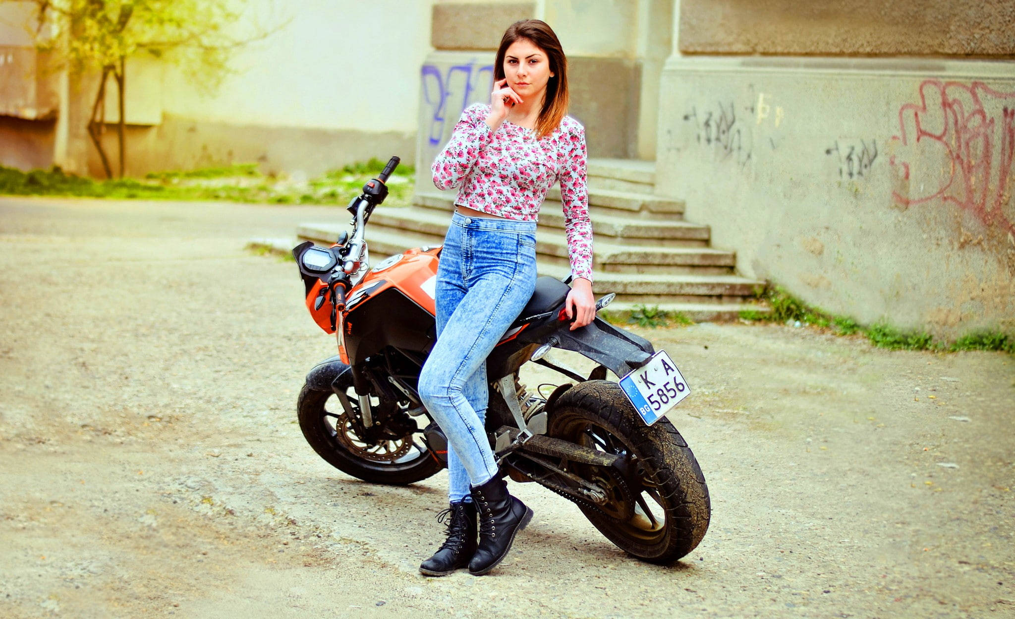 KTM 摩托車 桌布 壁紙 — 一位身穿花卉上衣和牛仔褲的年輕女性，隨意地靠在一輛停放在佈滿塗鴉的混凝土階梯旁的橙色機車上。