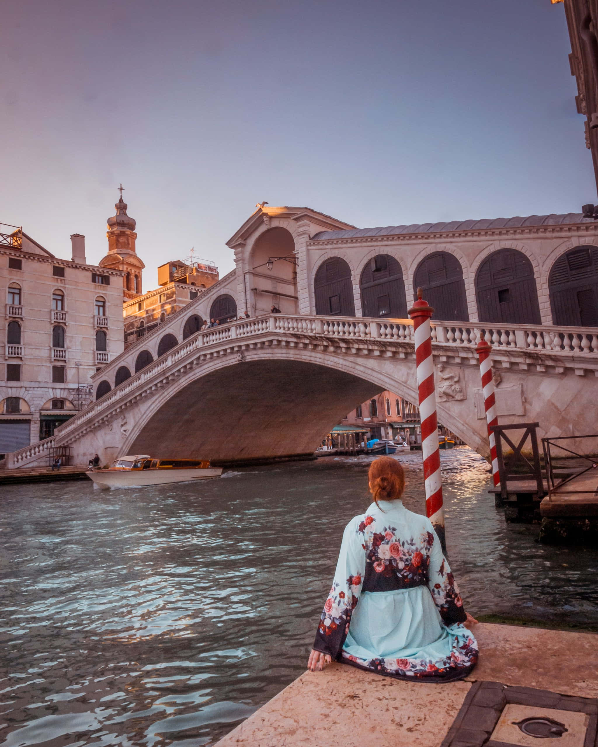 venice 桌布 壁紙 — 一位身穿花卉和服的人坐在威尼斯運河碼頭上，凝視著里亞爾托橋，一艘船在夕陽下從橋拱下滑過。