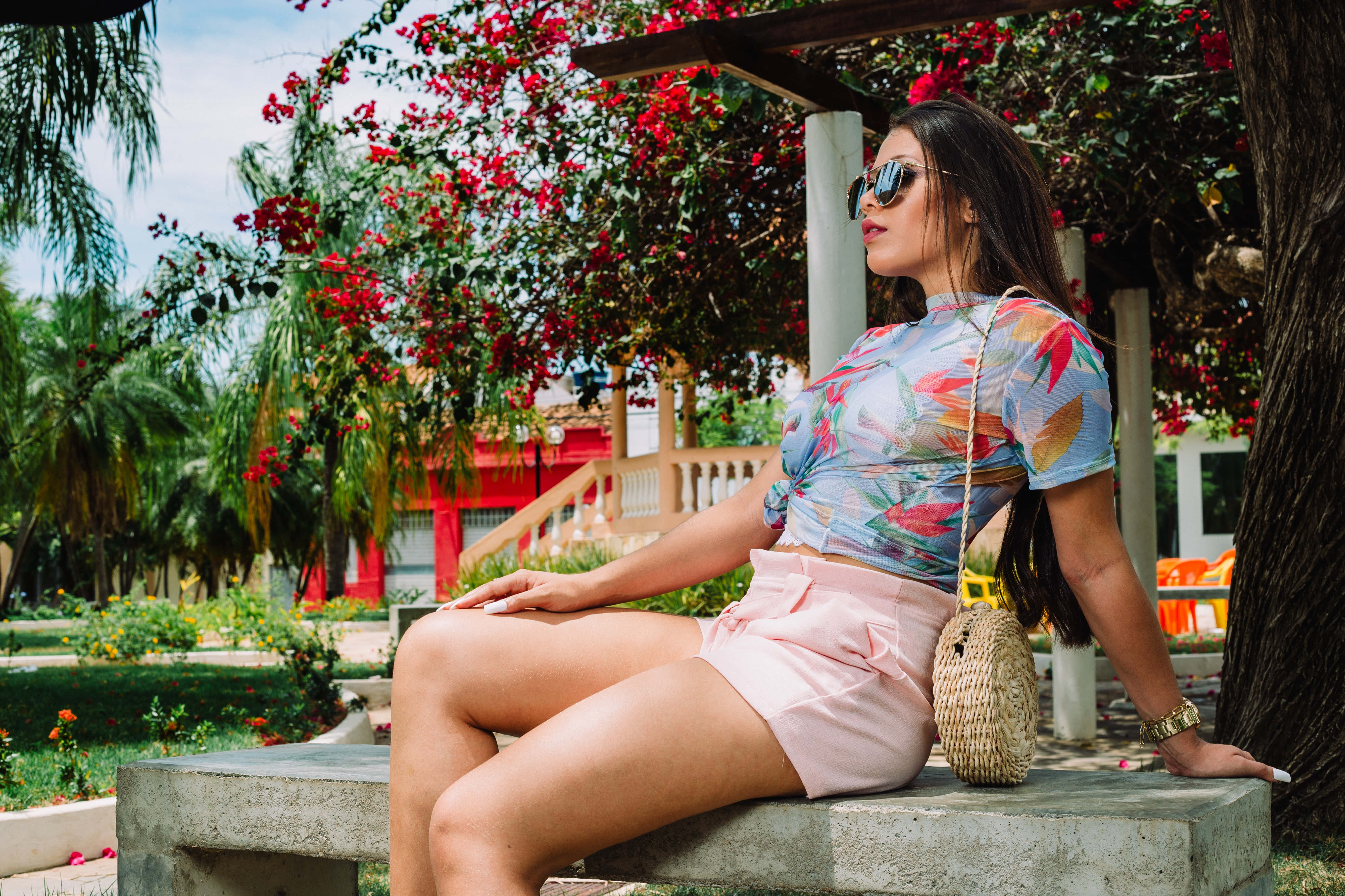 Summer Fashion 桌布 壁紙 — 一位身穿花卉圖案上衣和粉紅色短褲的女性，在盛開的紅花和棕櫚樹下的石凳上休息。