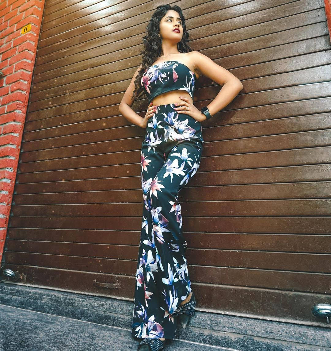 Nisha 桌布 壁紙 — 一位身穿花卉圖案兩件式套裝的女性自信地靠在木製百葉窗上，旁邊是磚牆。