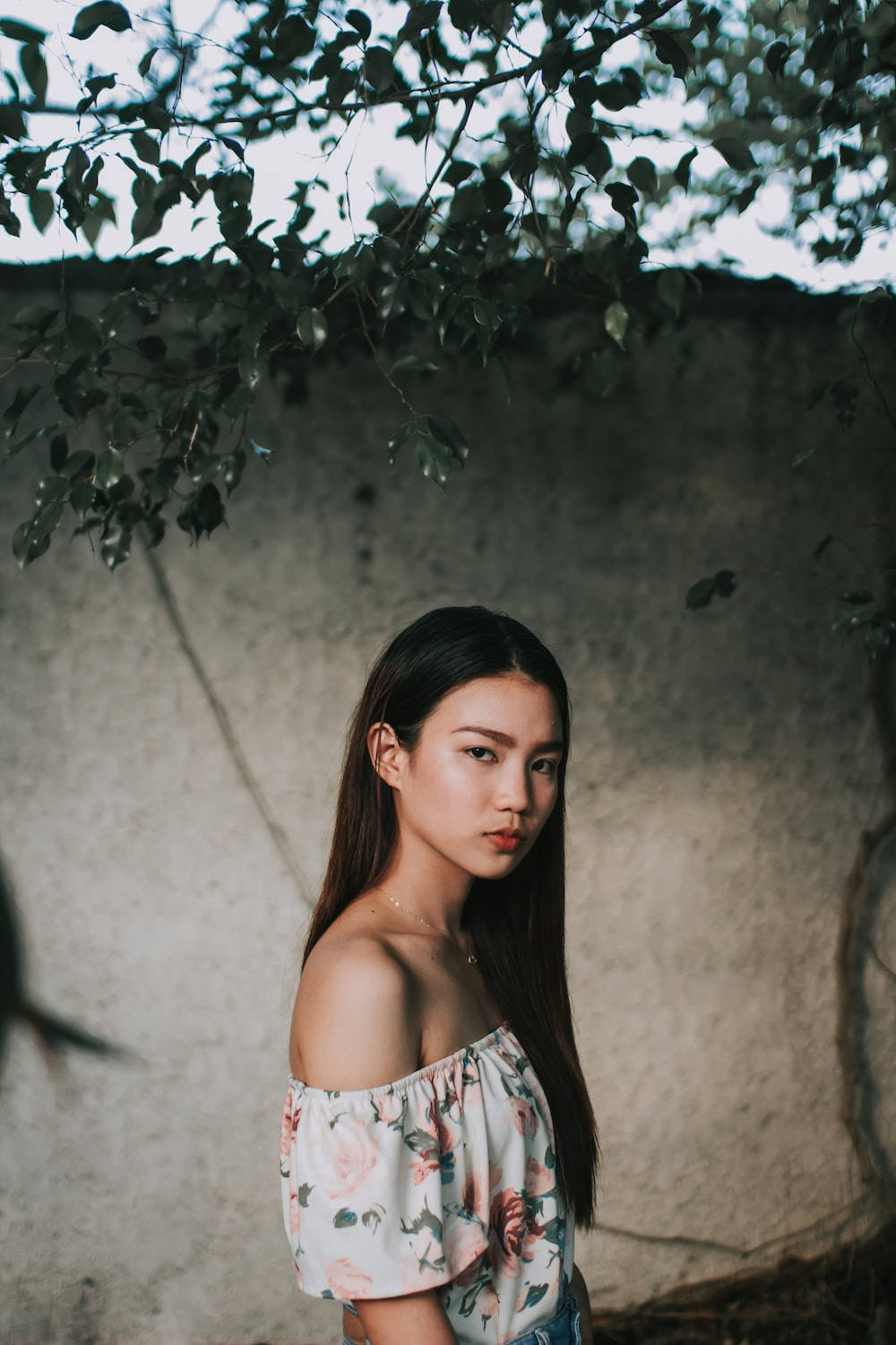 Thailand Woman 桌布 壁紙 — 一位身穿花卉圖案露肩上衣的年輕女性，站在枝葉茂密的樹枝下，背景是風化的混凝土牆。
