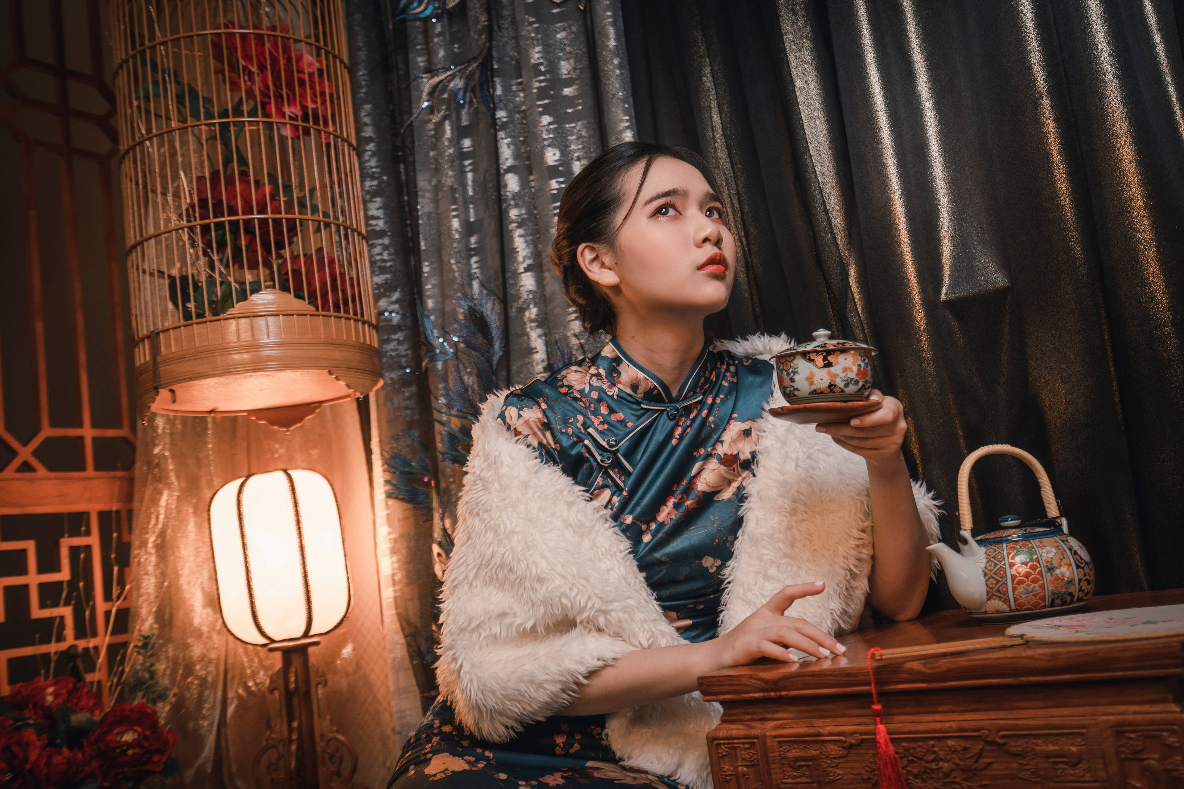 洋裝 桌布 壁紙 — 一位身穿花卉旗袍和白色毛皮披肩的女性，在溫暖的燈籠光下，拿著茶杯，旁邊是一個華麗的茶壺。