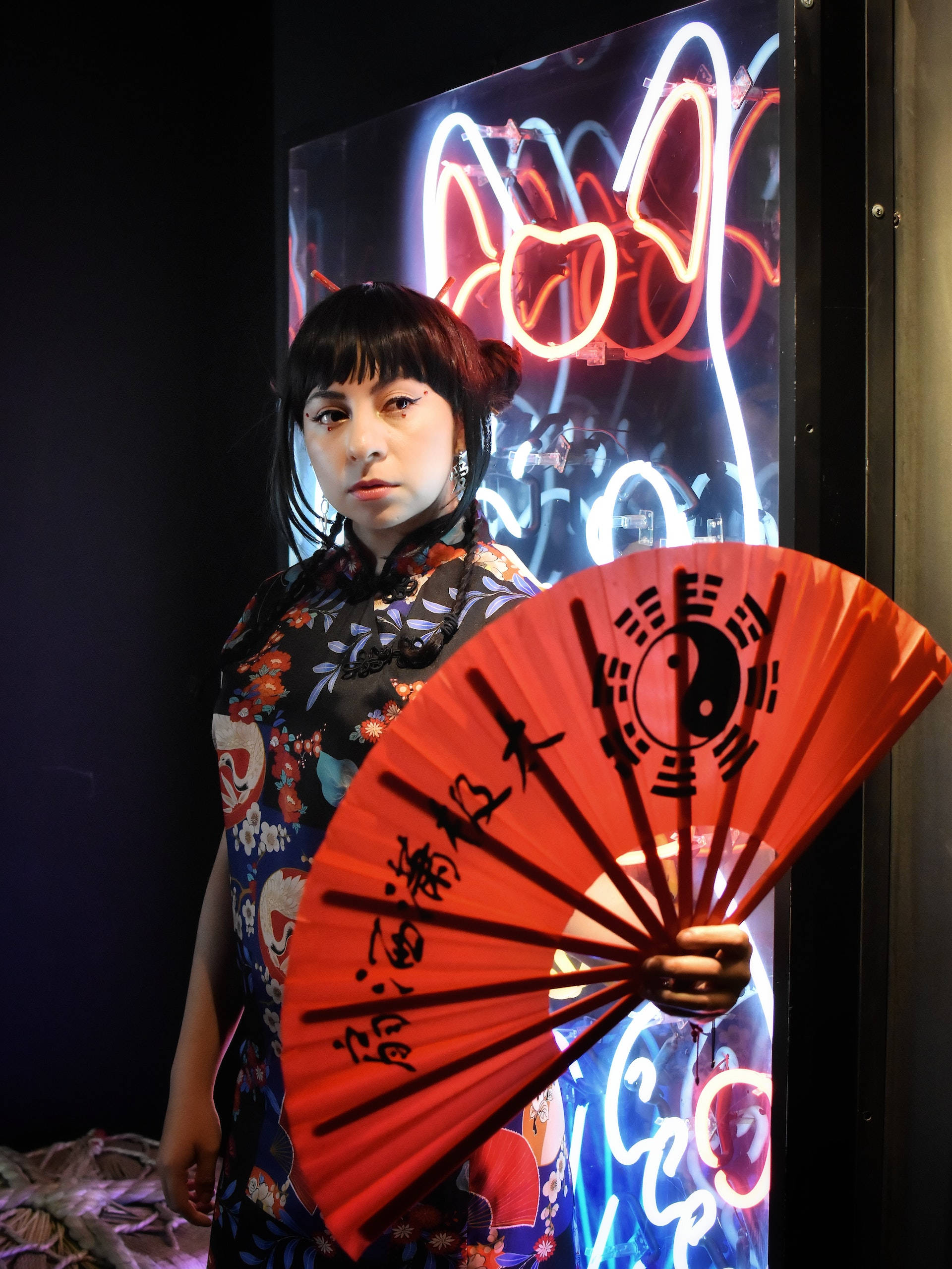 Asian Woman 桌布 壁紙 — 一位身穿花卉旗袍的女性，在閃爍的霓虹燈招牌背景下，手持帶有陰陽圖案的紅色扇子。