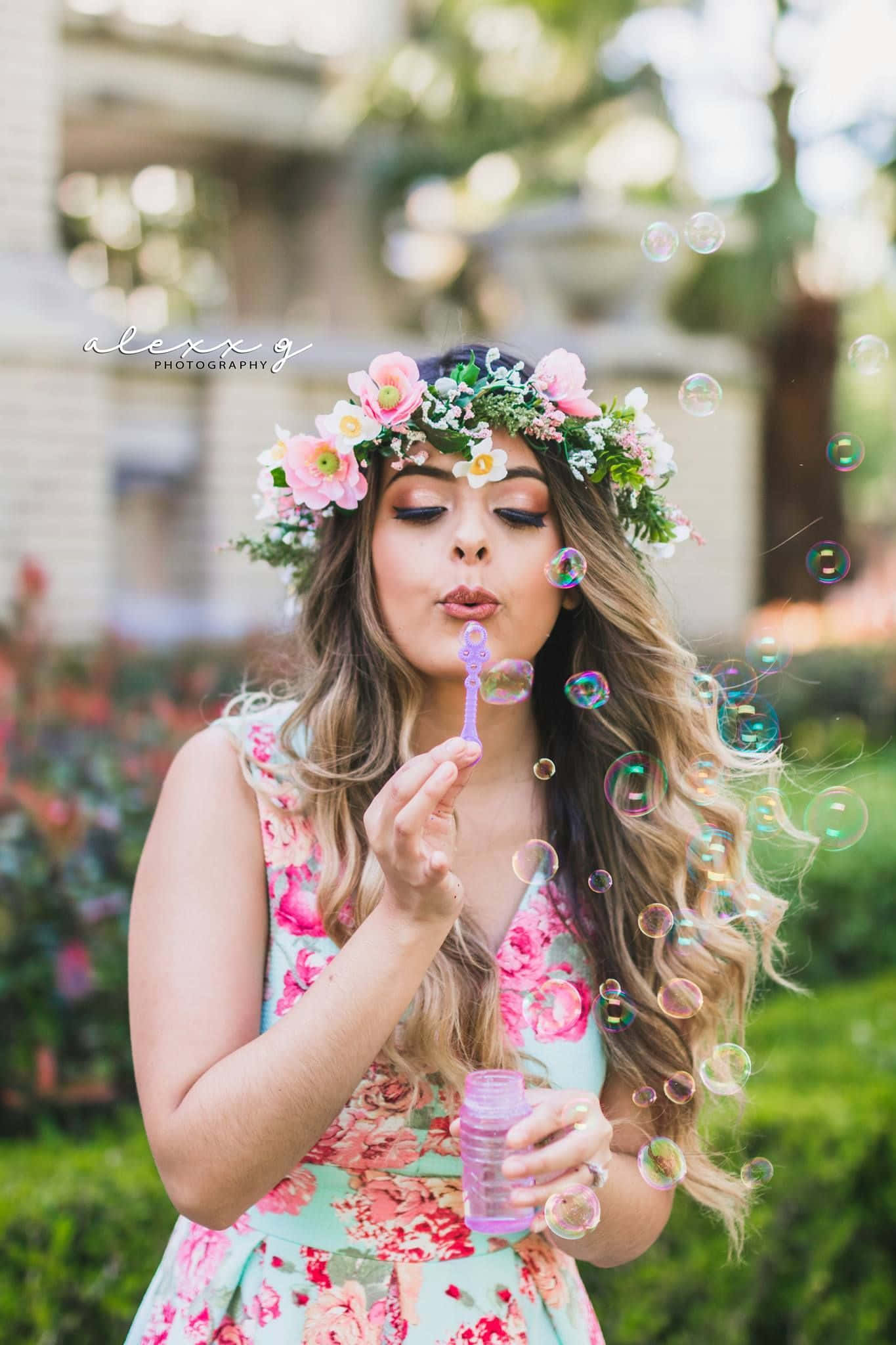 Floral Crown 桌布 壁紙 — 一位身穿花卉洋裝、頭戴皇冠的年輕女子，在陽光普照的花園中吹出色彩繽紛的泡泡，背景帶有柔和散景效果。
