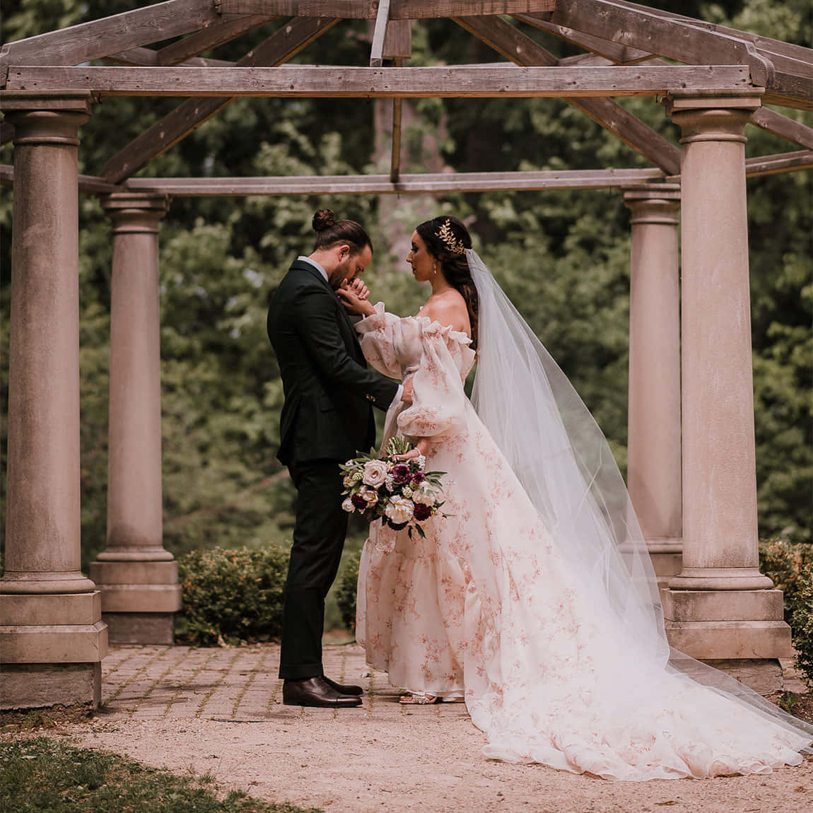 Garden Wedding 桌布 壁紙 — 一位身穿花卉禮服和頭紗的新娘，在石亭下輕撫新郎的臉龐，四周環繞著茂密的綠植。