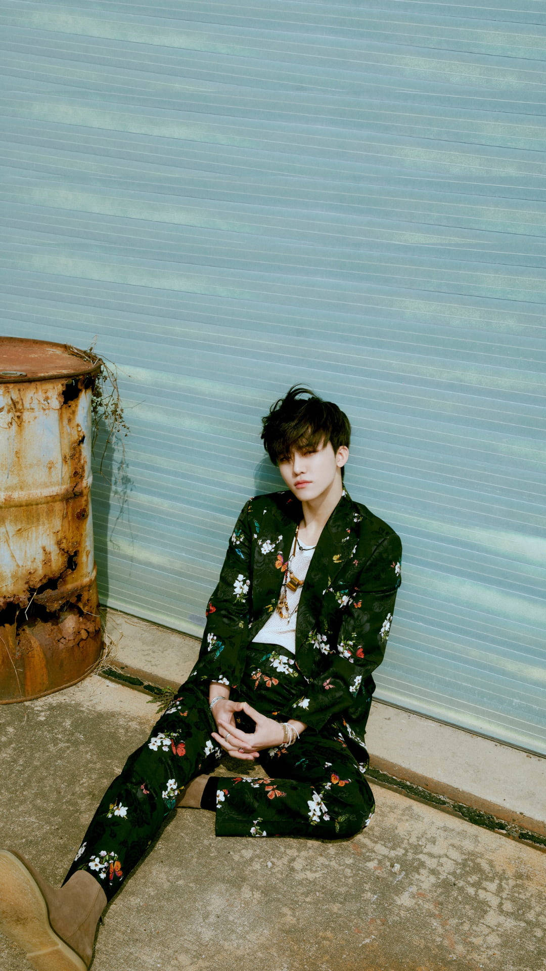 jaemin 桌布 壁紙 — 一位身穿花卉綠色西裝的年輕男子，靠著一個風化的藍色金屬百葉窗，旁邊有一個生鏽的桶子，若有所思地坐著。