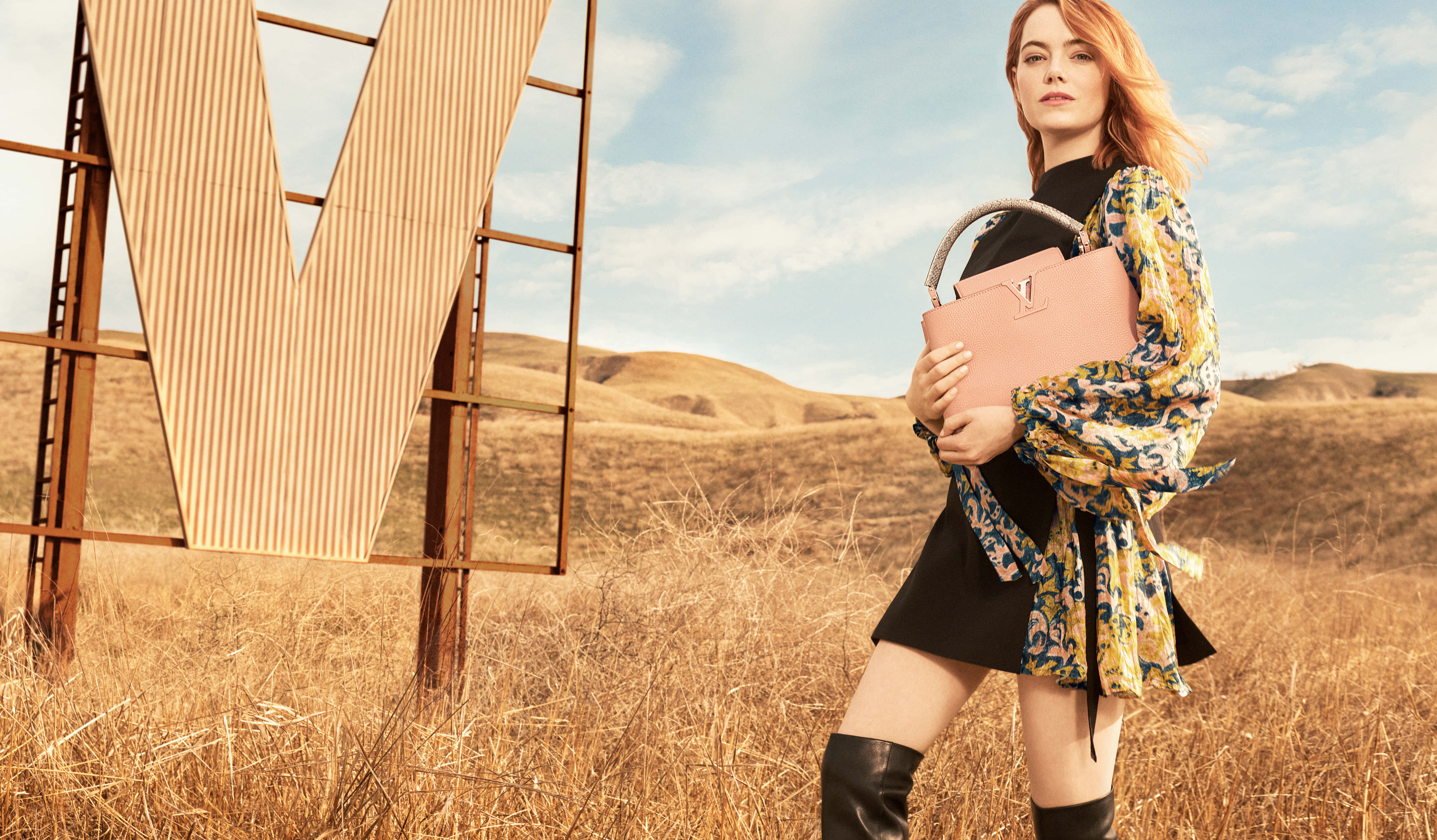 Luxury Brands 桌布 壁紙 — 一位身穿花卉襯衫和黑色靴子的女性，自信地站在乾燥草地上一個巨大的生鏽金屬V標誌旁，背景是明亮的藍天。