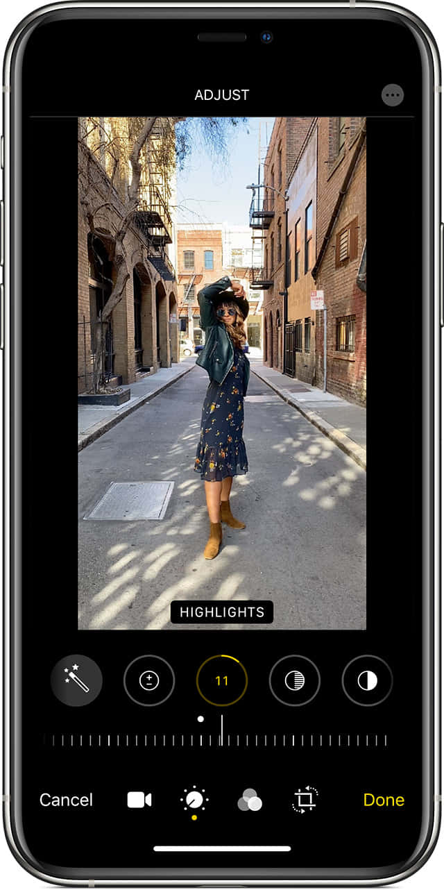 iPhone 桌布 壁紙 — 一位身穿花卉連衣裙和靴子的女性在磚砌建築之間的陽光普照的城市小巷中俏皮地擺姿勢。