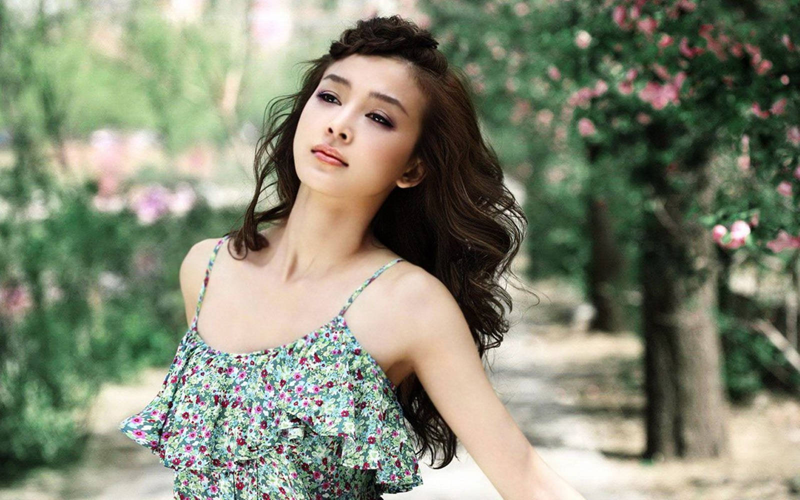 美女 桌布 壁紙 — 一位身穿花卉連衣裙的年輕女子優雅地站在陽光普照的花園中，周圍是盛開的粉紅色樹木。