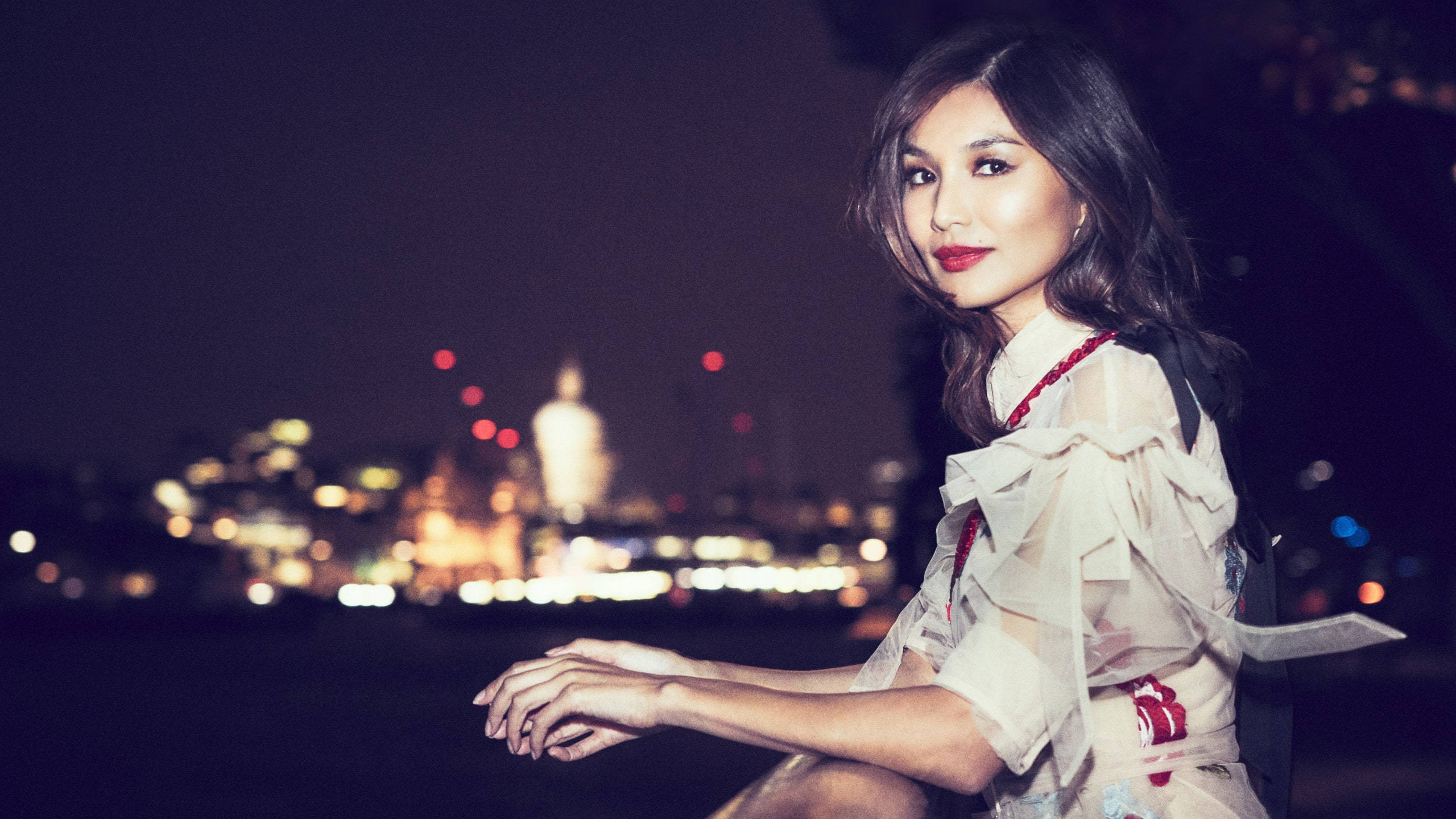 Gemma Chan 桌布 壁紙 — 一位身穿荷葉邊白色連衣裙的女性在夜晚坐在水邊，身後是散景的城市景觀閃爍著光芒。