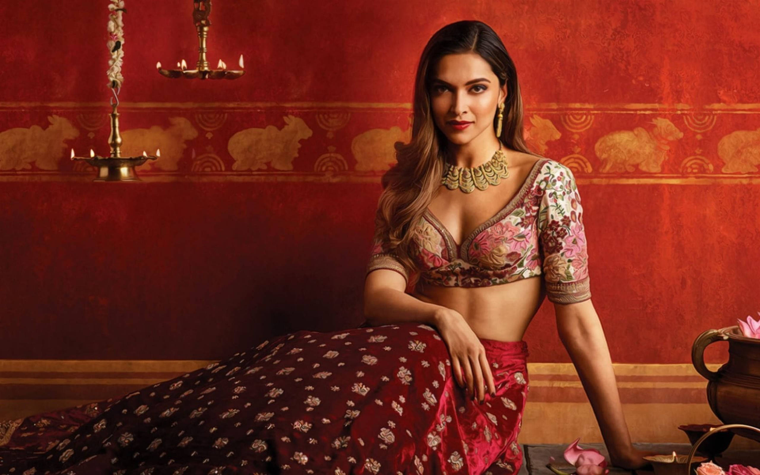 Deepika 桌布 壁紙 — 一位身穿華麗傳統印度服飾的女性，在帶有金色兔子圖案和懸掛油燈的豐富紅色紋理牆壁前優雅地擺姿勢。