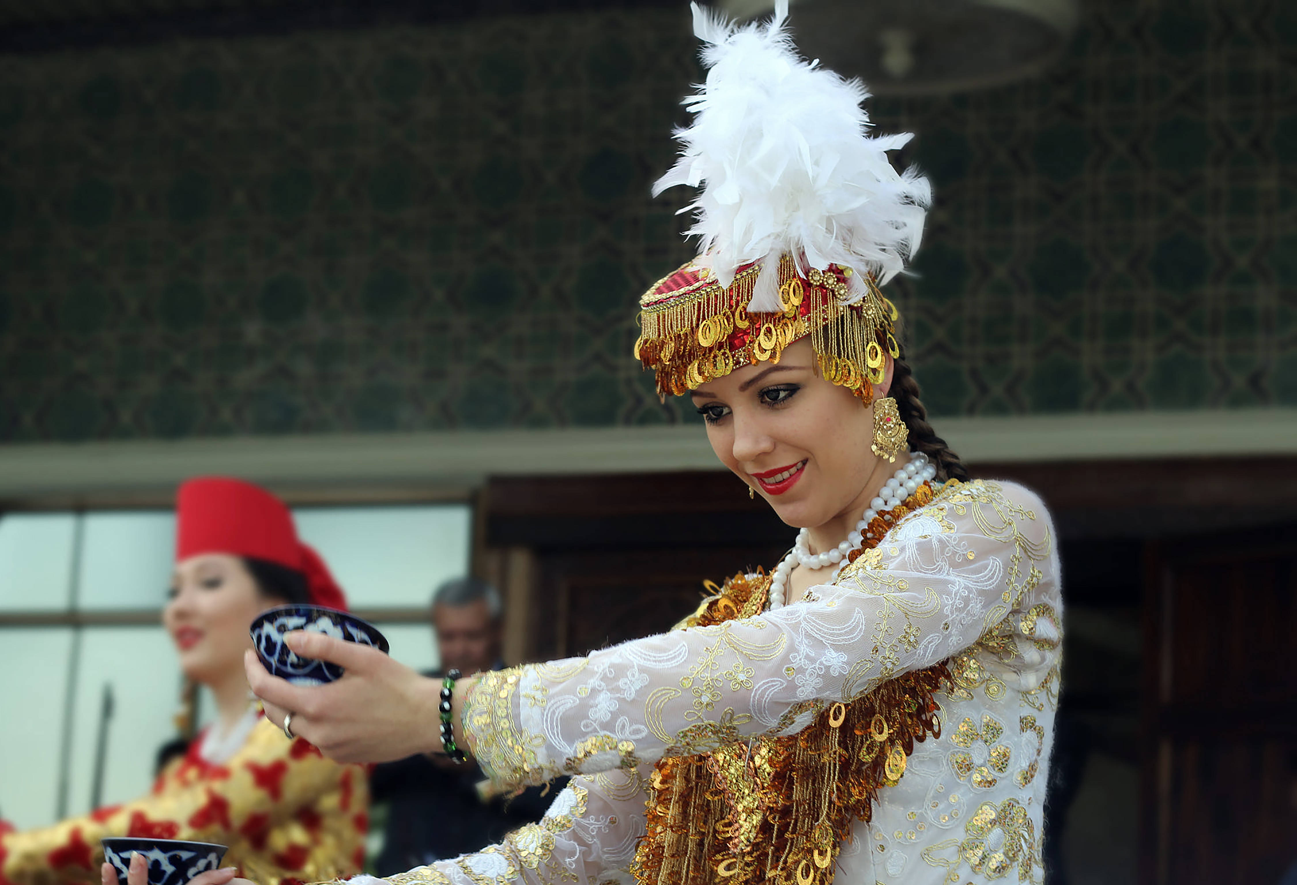 Uzbekistan 桌布 壁紙 — 一位身穿華麗傳統服飾、頭戴羽毛頭飾的女性，優雅地從一個有圖案的碗中倒出東西。