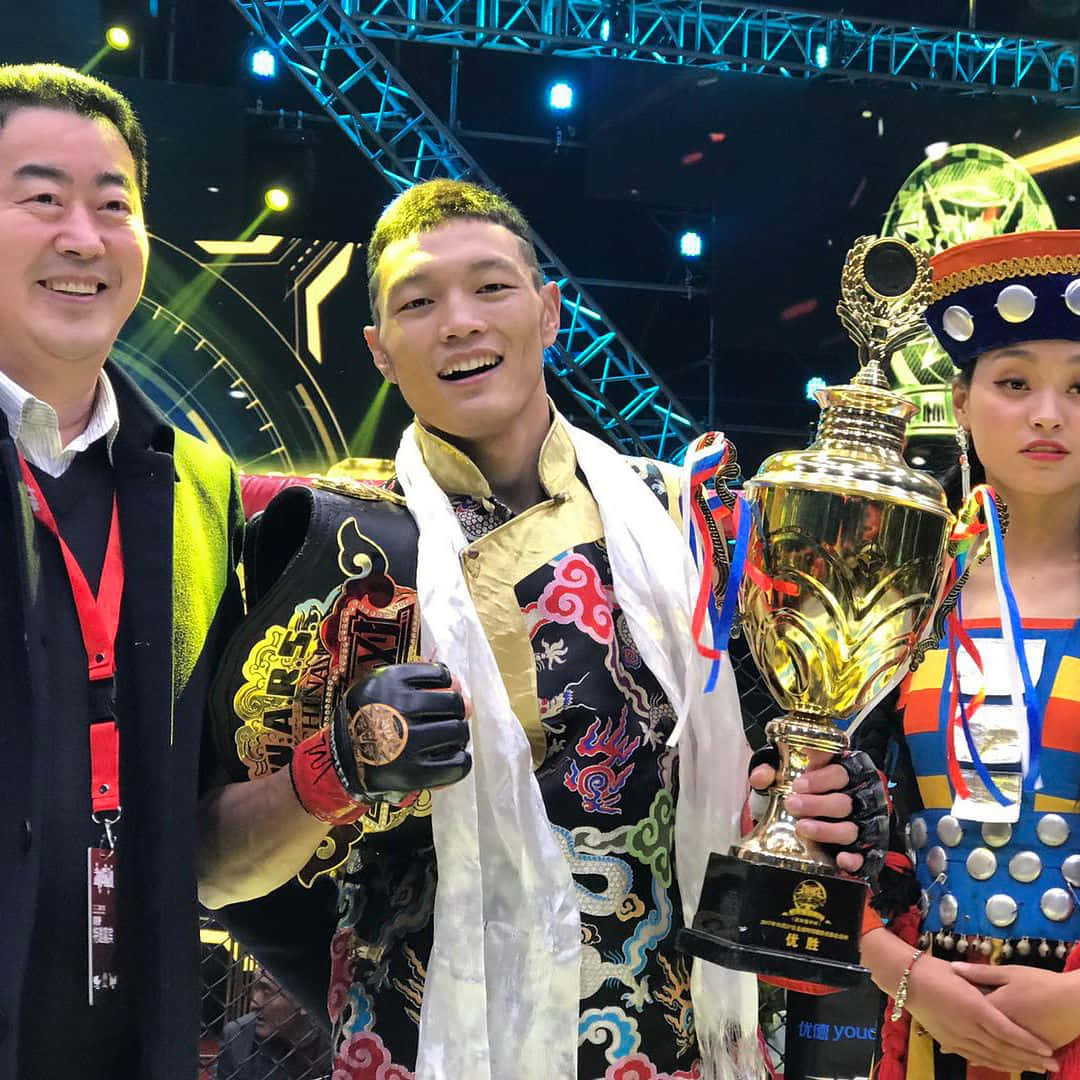 MMA 桌布 壁紙 — 一位身穿華麗傳統服飾的勝利戰士，在競技場燈光下，手持冠軍獎盃，身旁有兩位微笑的同伴，滿臉光彩。