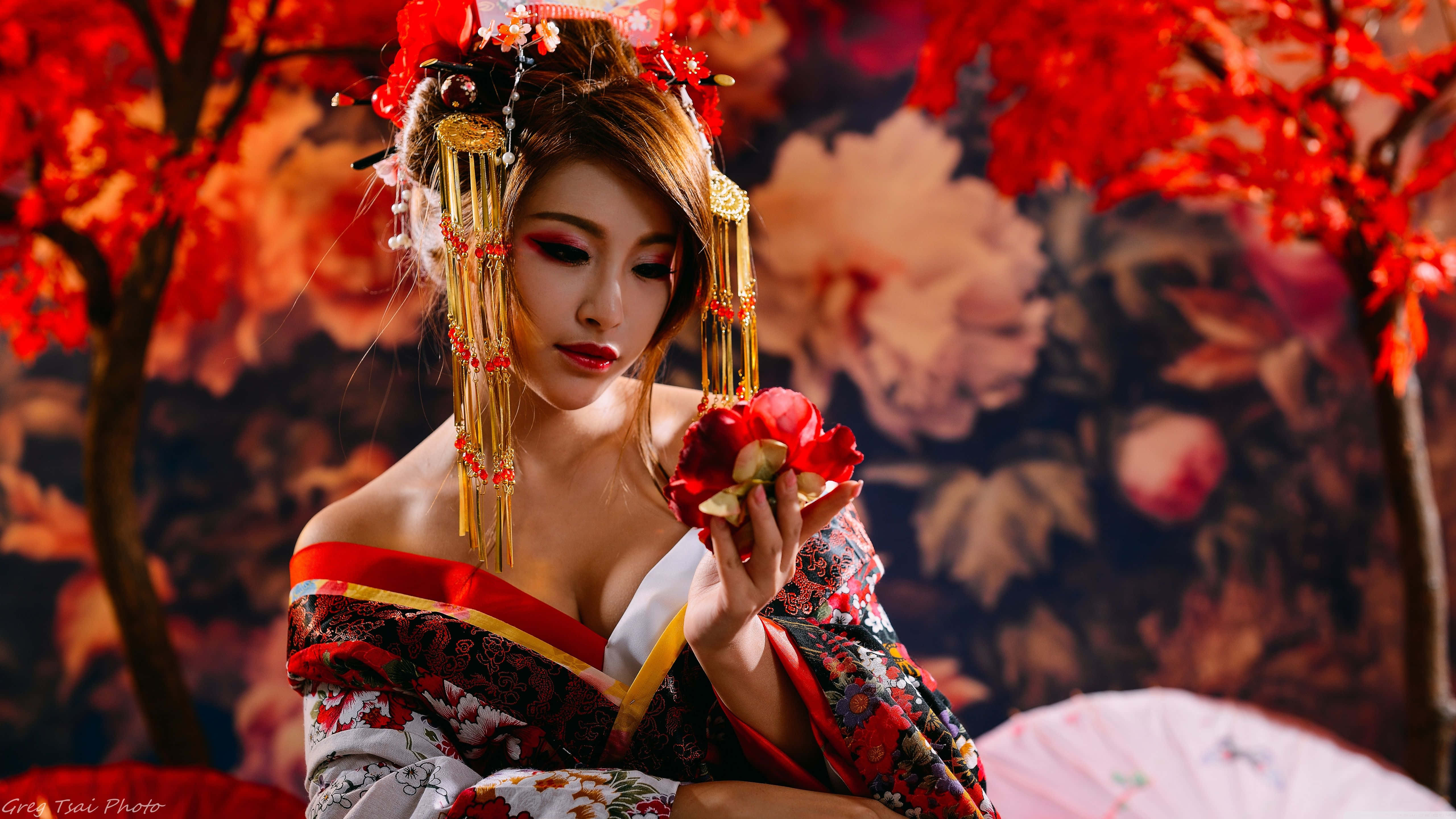 Japanese Women 桌布 壁紙 — 一位身穿華麗傳統服飾的女子，在熾熱的秋葉和花卉圖案背景下手持一朵紅花。