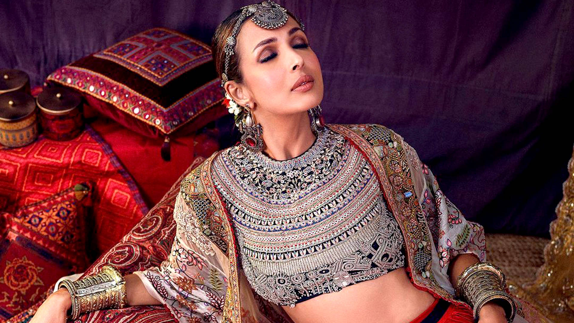 Bollywood HD 桌布 壁紙 — 一位身穿華麗傳統服飾的女性，在圖案豐富的紡織品和裝飾靠墊中優雅地斜倚著。