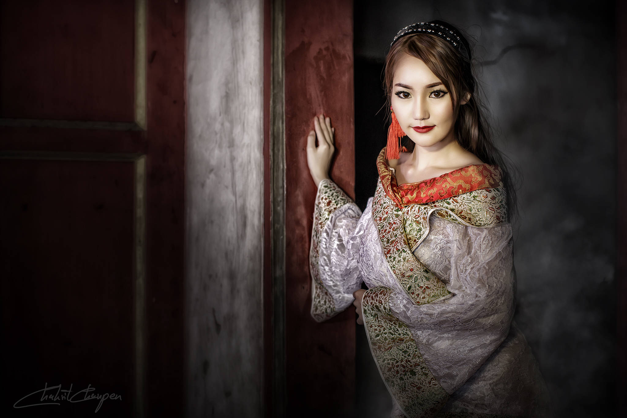 Thailand Woman 桌布 壁紙 — 一位身穿華麗傳統服飾的女性，在昏暗且充滿氛圍的環境中，從一扇風化的木門後探出頭來。