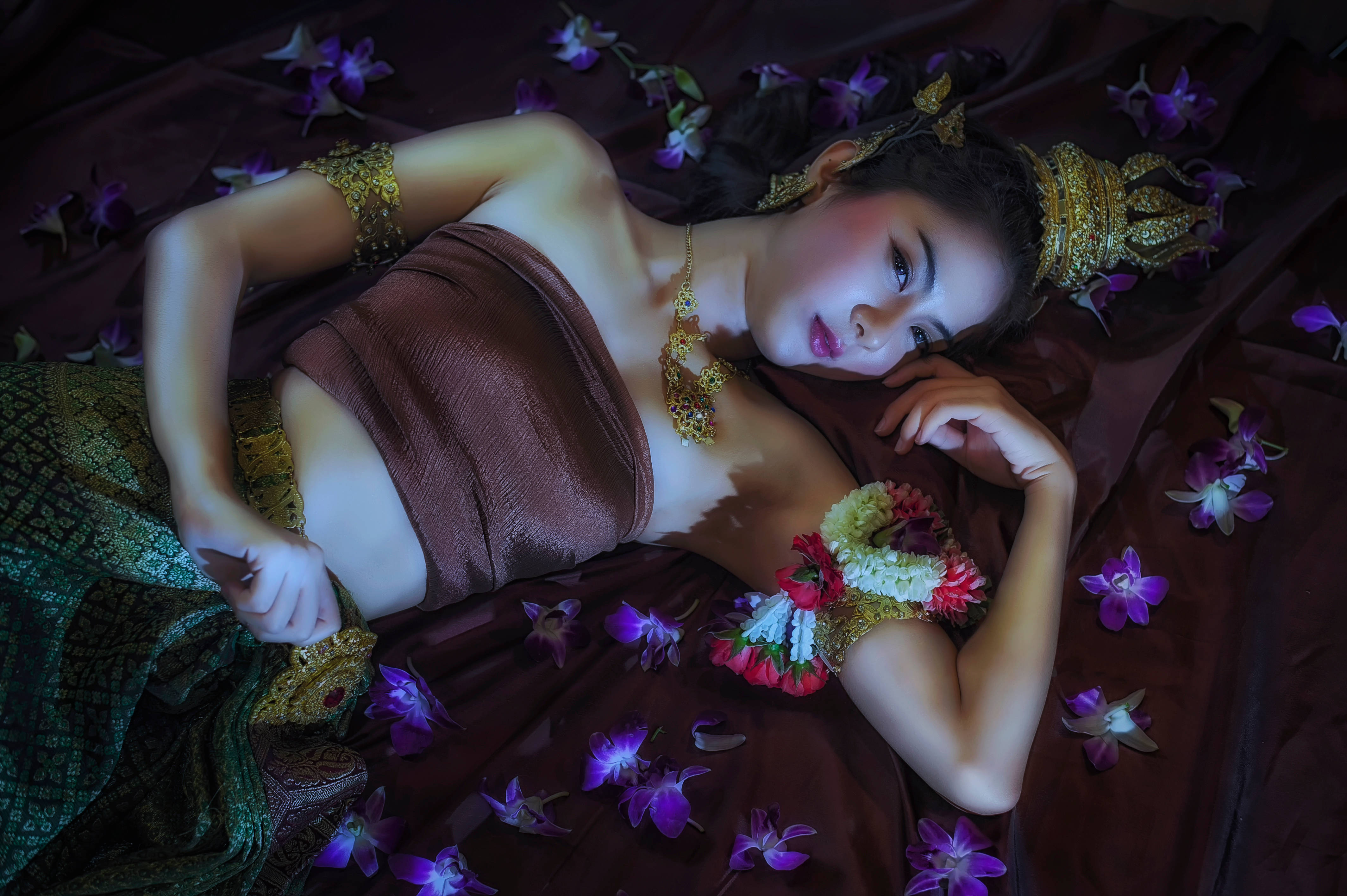Thailand Woman 桌布 壁紙 — 一位身穿華麗傳統服飾的女性，在柔和燈光下優雅地躺在深栗色布料上的紫色蘭花叢中。