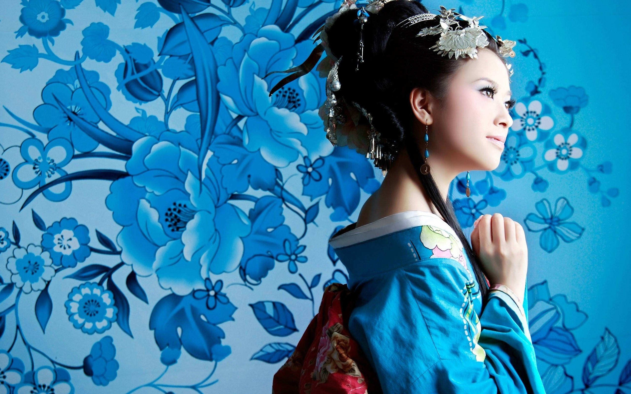 Geisha 桌布 壁紙 — 一位身穿華麗傳統服飾的女性，在鮮豔的藍色花卉壁紙背景前優雅地站立。