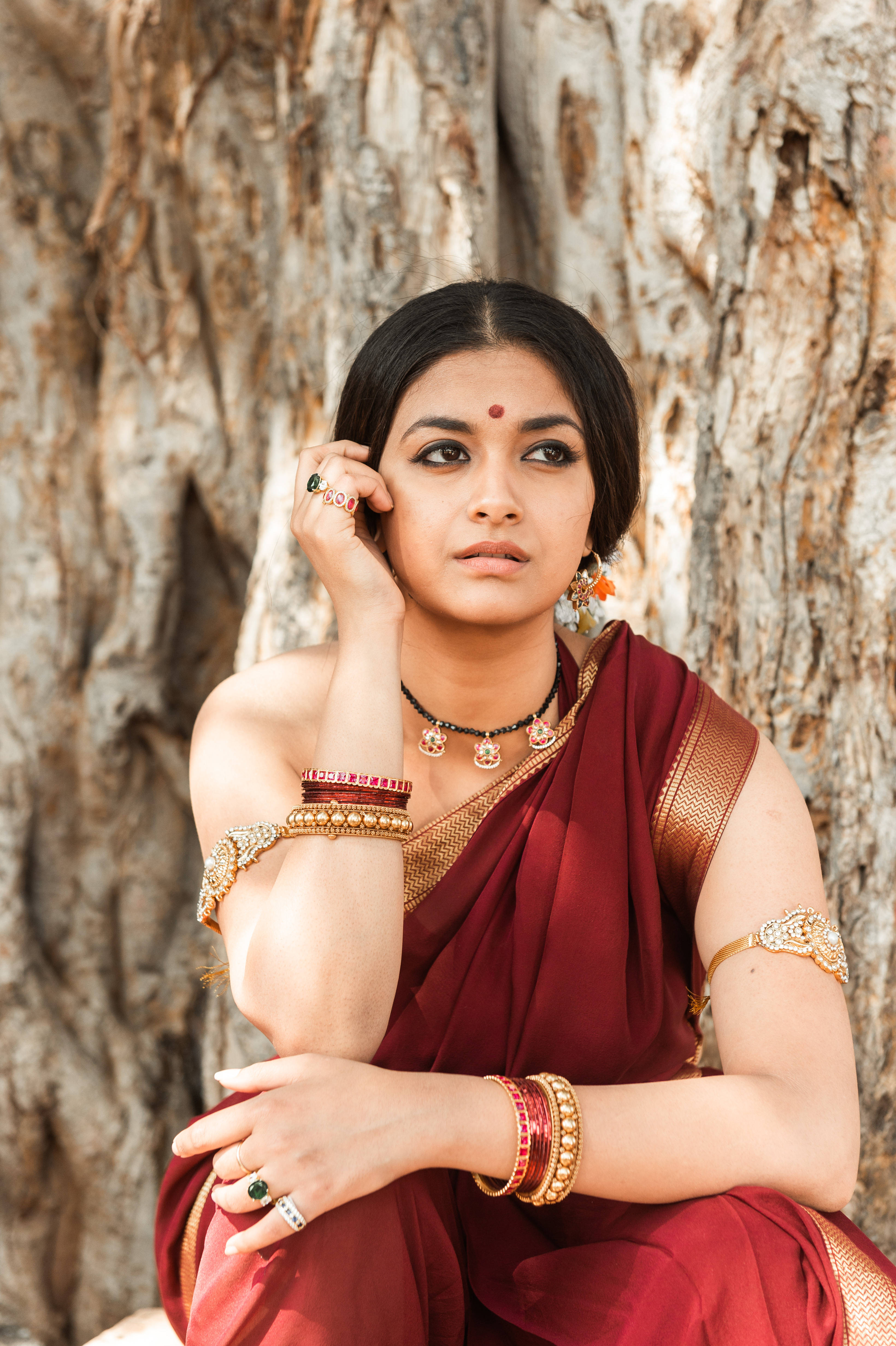 Indian 桌布 壁紙 — 一位身穿華麗栗色紗麗、佩戴金飾的女子，在紋理樹幹前優雅地擺姿勢。
