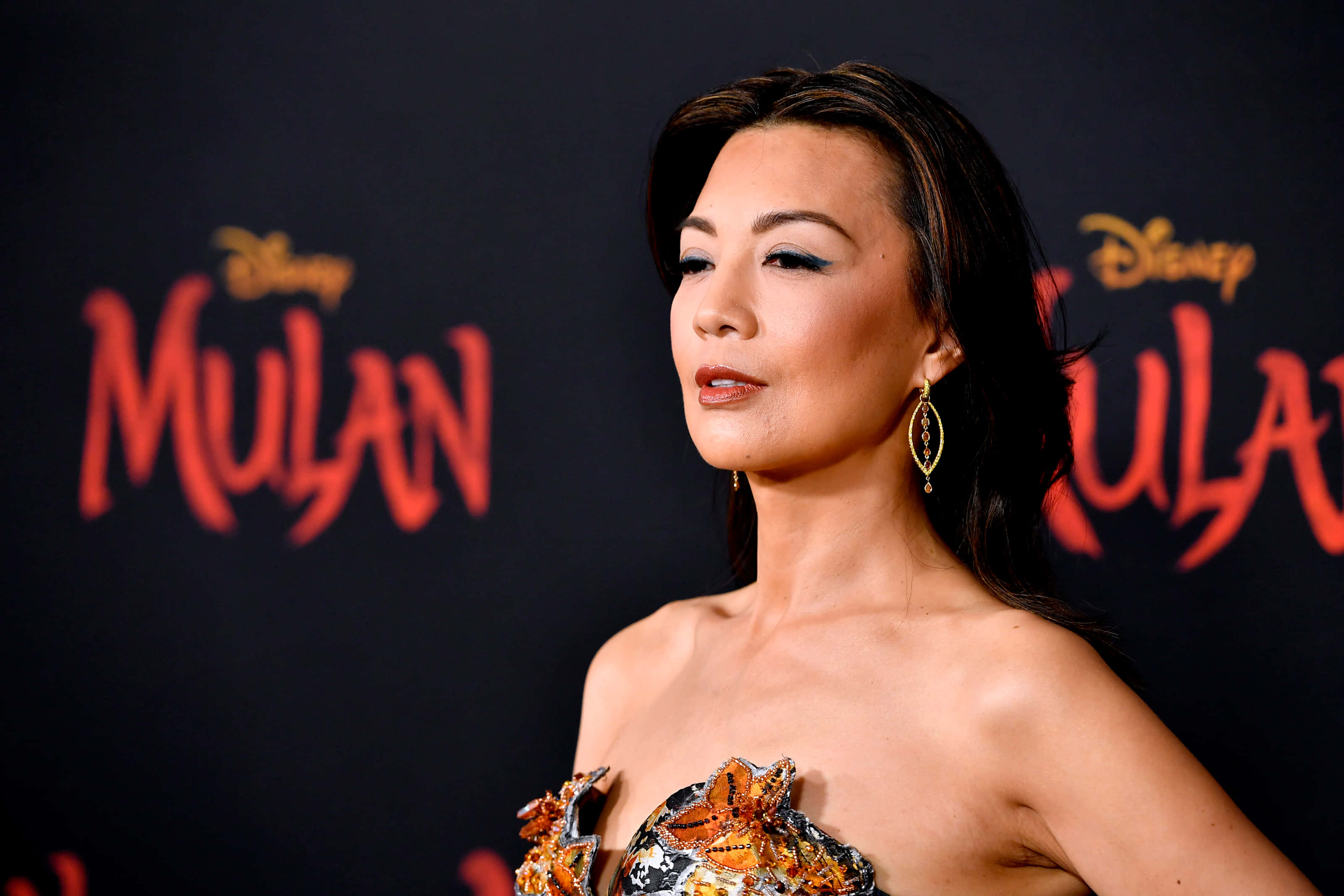 Ming Na Wen 桌布 壁紙 — 一位身穿華麗無肩帶禮服的女士在迪士尼花木蘭首映會上，以醒目的紅色標誌背景擺姿勢。