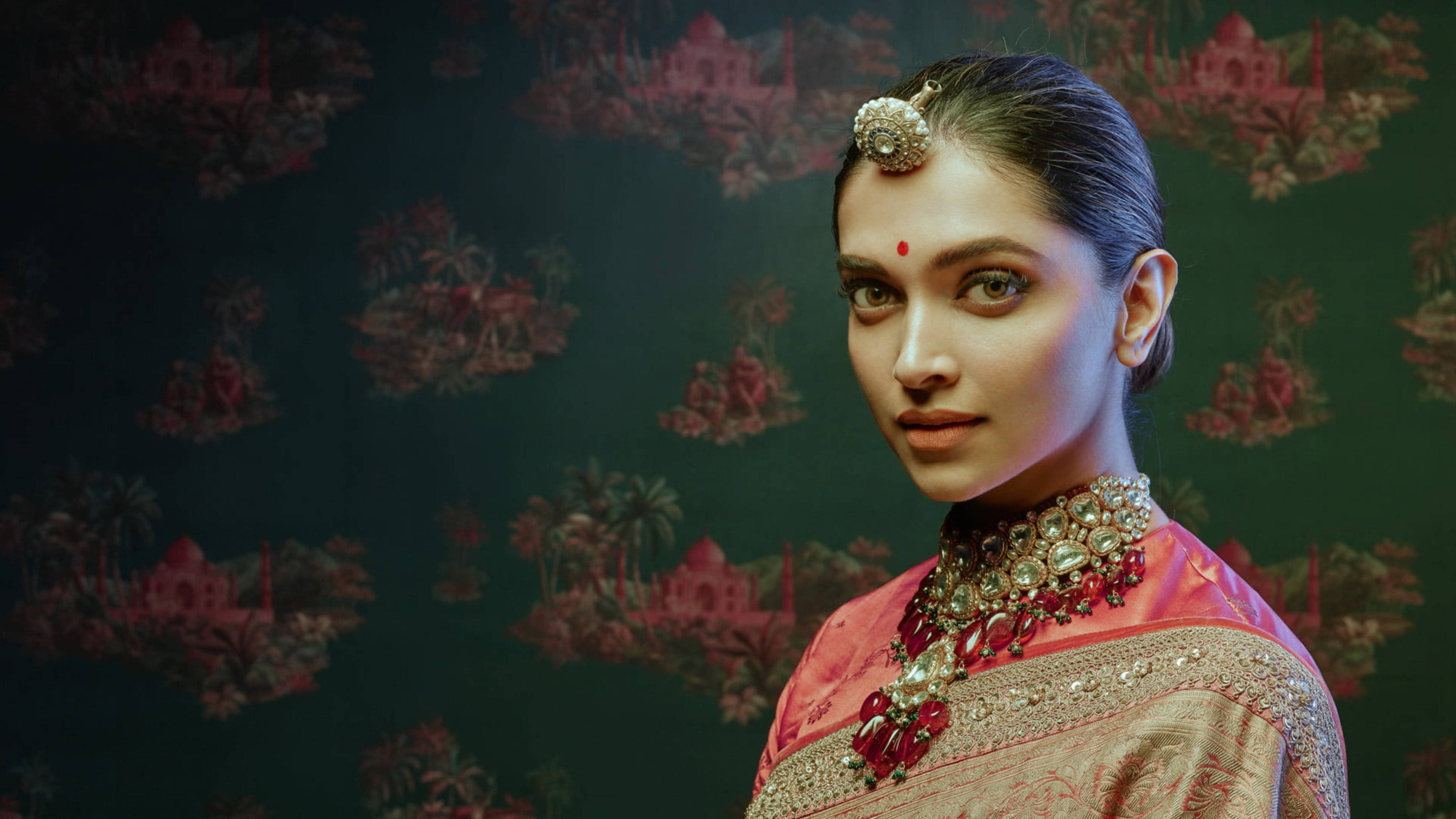 Deepika 桌布 壁紙 — 一位身穿華麗粉紅金色傳統服飾、額頭點有紅色吉祥痣、頭戴珠寶頭飾的女性，站在深綠色帶有粉紅泰姬陵圖案的壁紙前。
