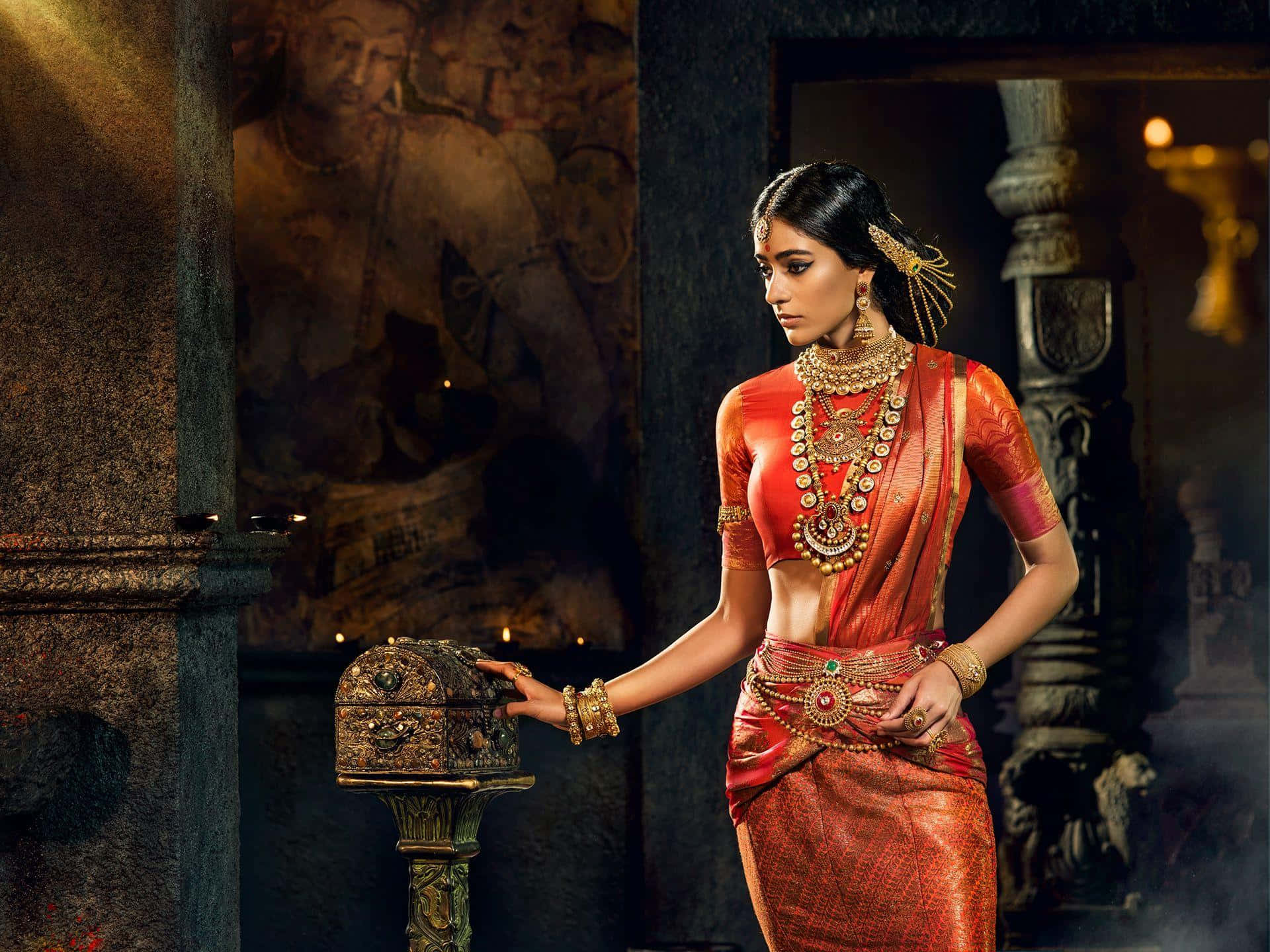 Indian 桌布 壁紙 — 一位身穿華麗紅色傳統服飾、佩戴金飾的女子，站在燈光昏暗的古老寺廟中，觸摸一個雕刻精美的箱子。