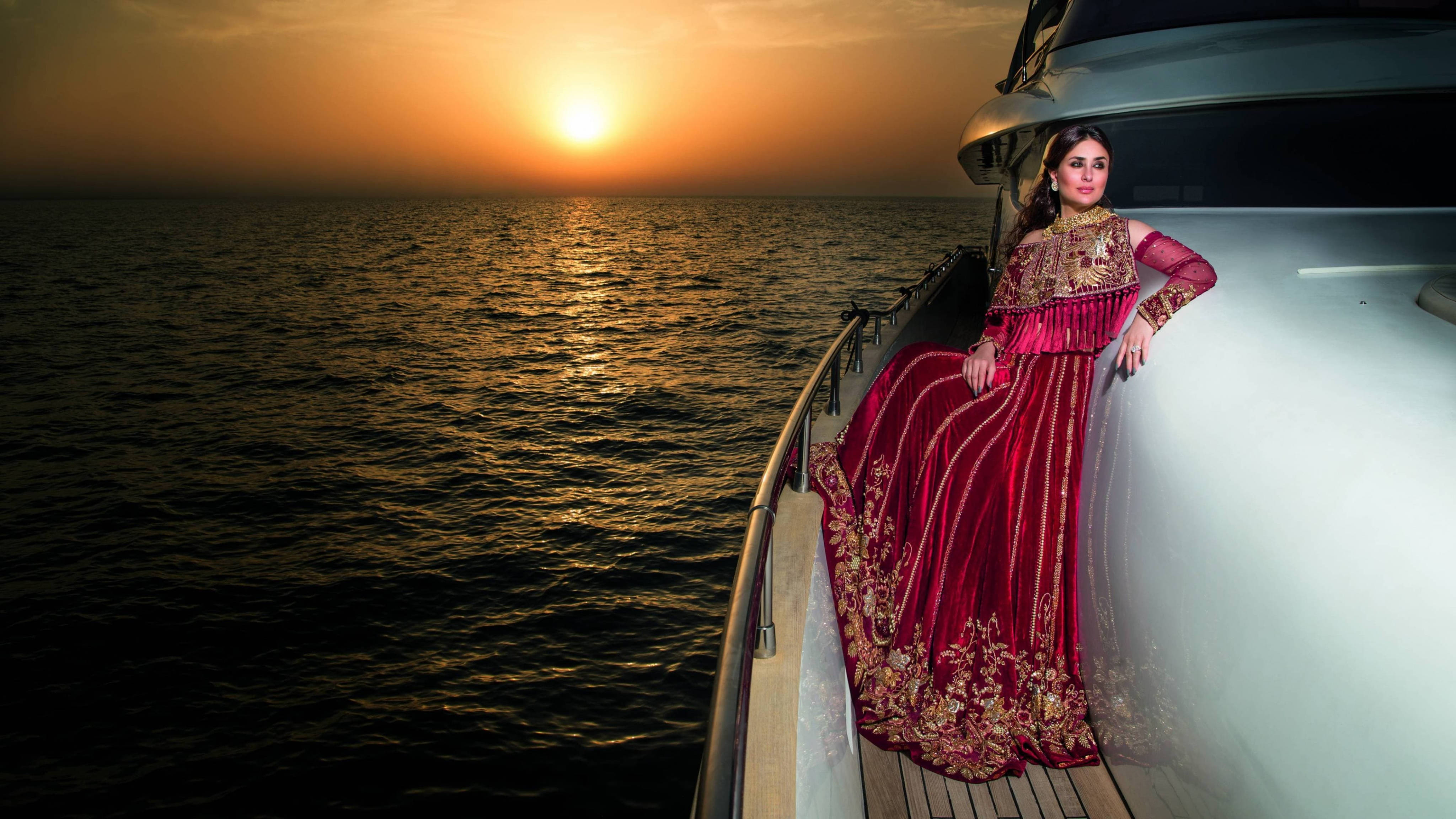 Kareena 桌布 壁紙 — 一位身穿華麗紅色刺繡長袍的女性，在夕陽西下平靜的海面上，優雅地站在遊艇甲板上。
