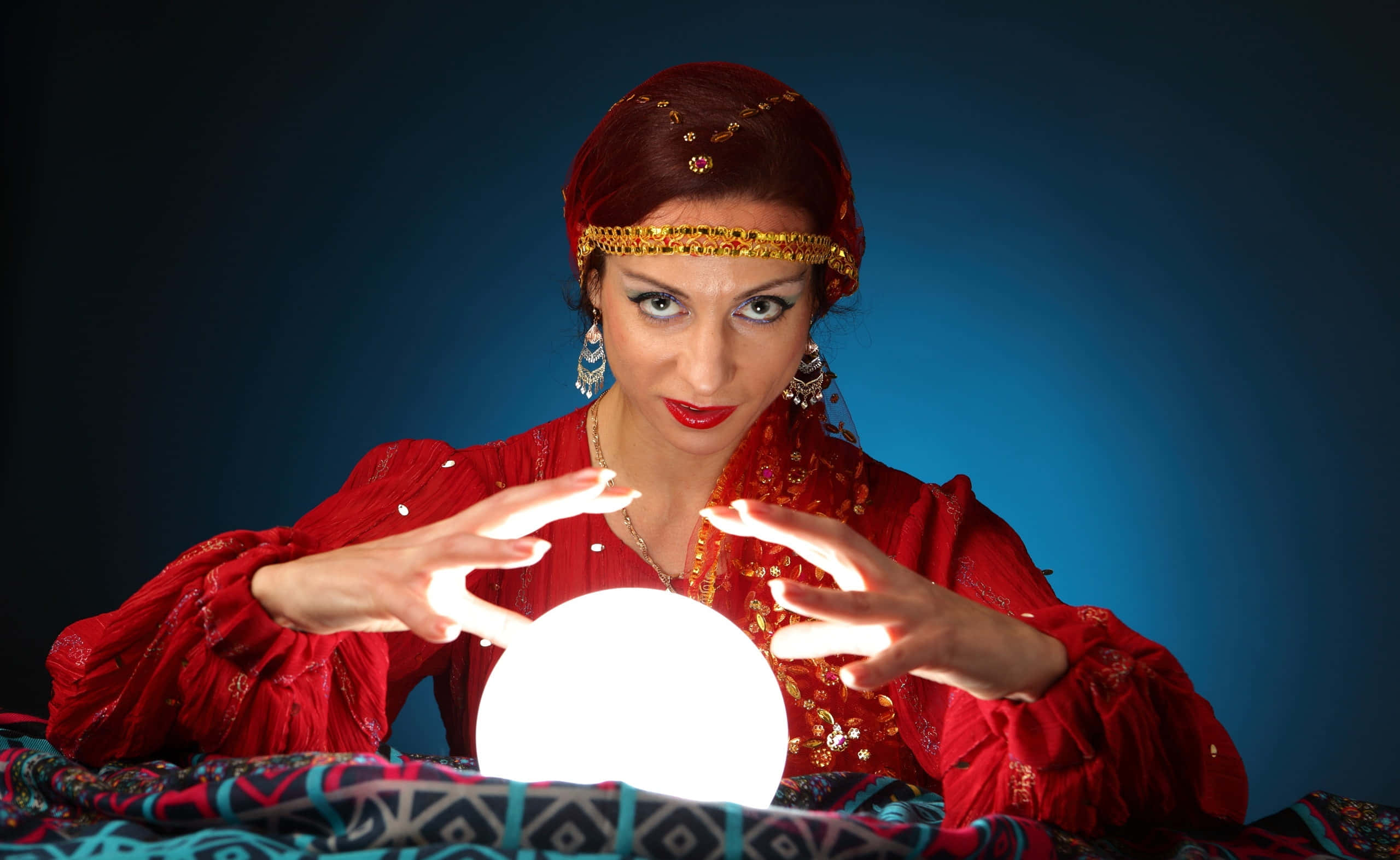 Fortune Teller 桌布 壁紙 — 一位身穿華麗紅色服裝的女性，雙手懸浮在發光的水晶球上方，專注地凝視著它，背景是深藍色漸變。