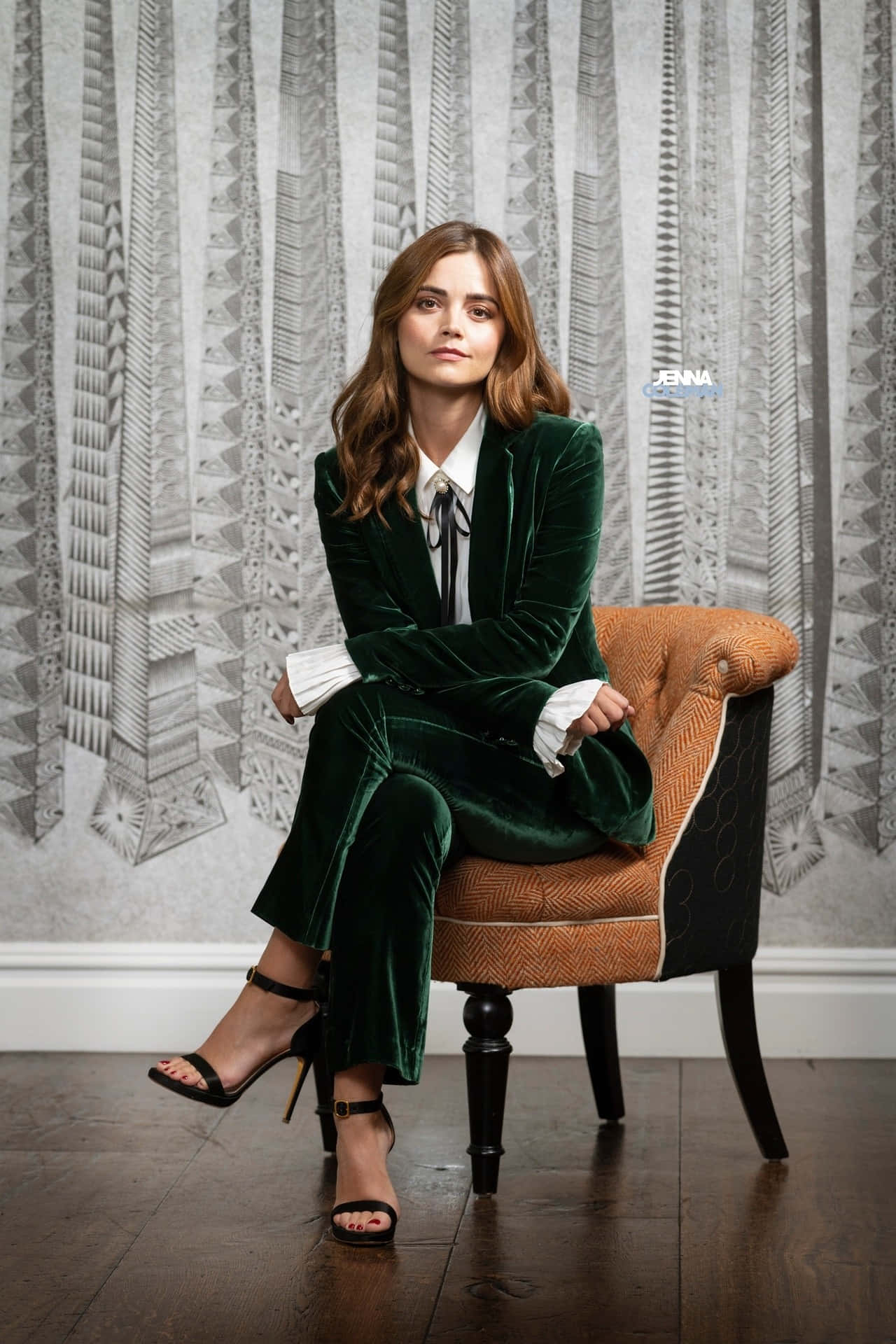 Jenna Coleman 桌布 壁紙 — 一位身穿華麗綠色天鵝絨西裝的女性，端坐在圖案扶手椅上，背景是幾何圖案壁紙。