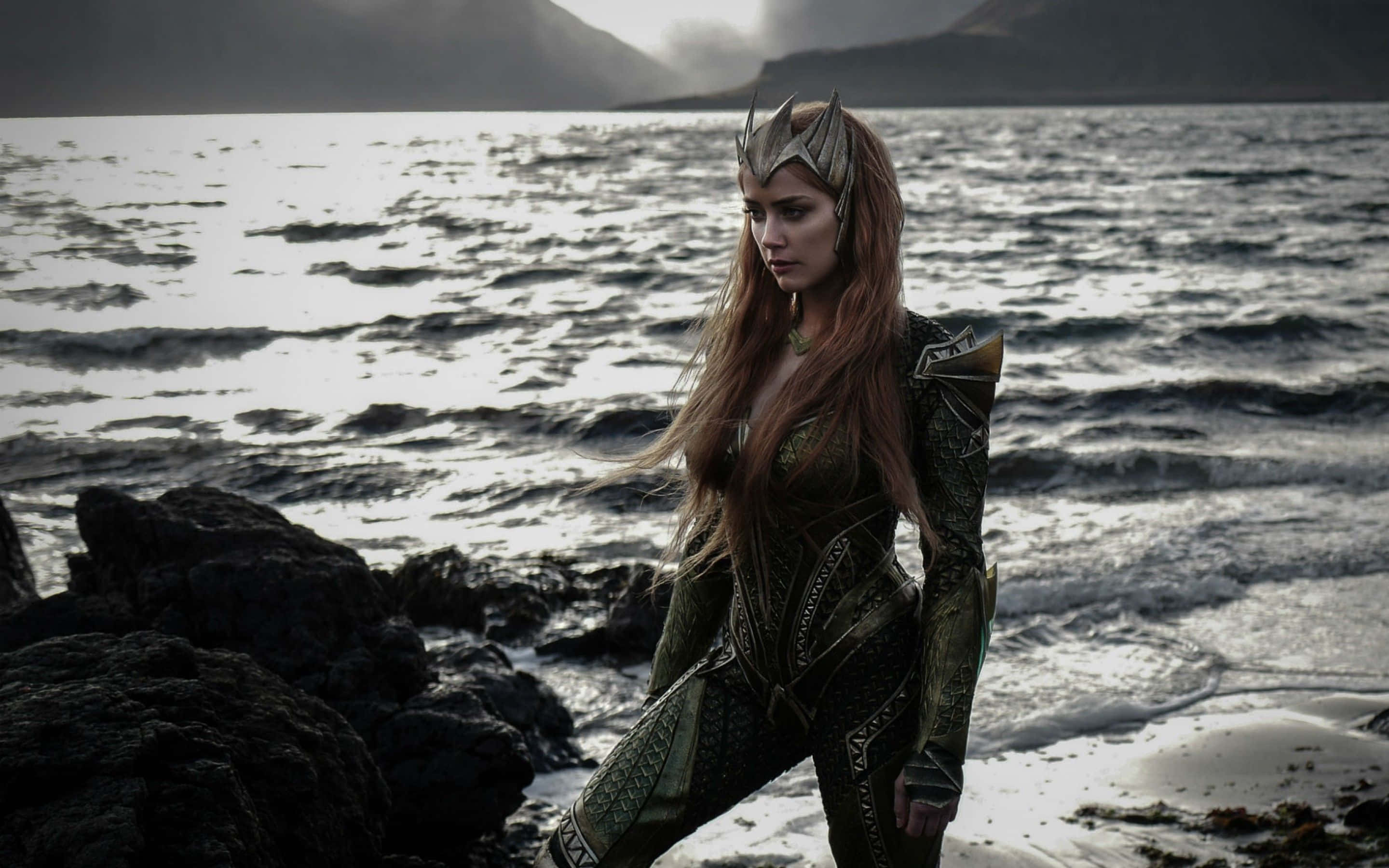 Amber Heard 桌布 壁紙 — 一位身穿華麗綠色鎧甲的女戰士女王站在岩石海岸線上，波濤在陰沉天空下拍打著暗色岩石。