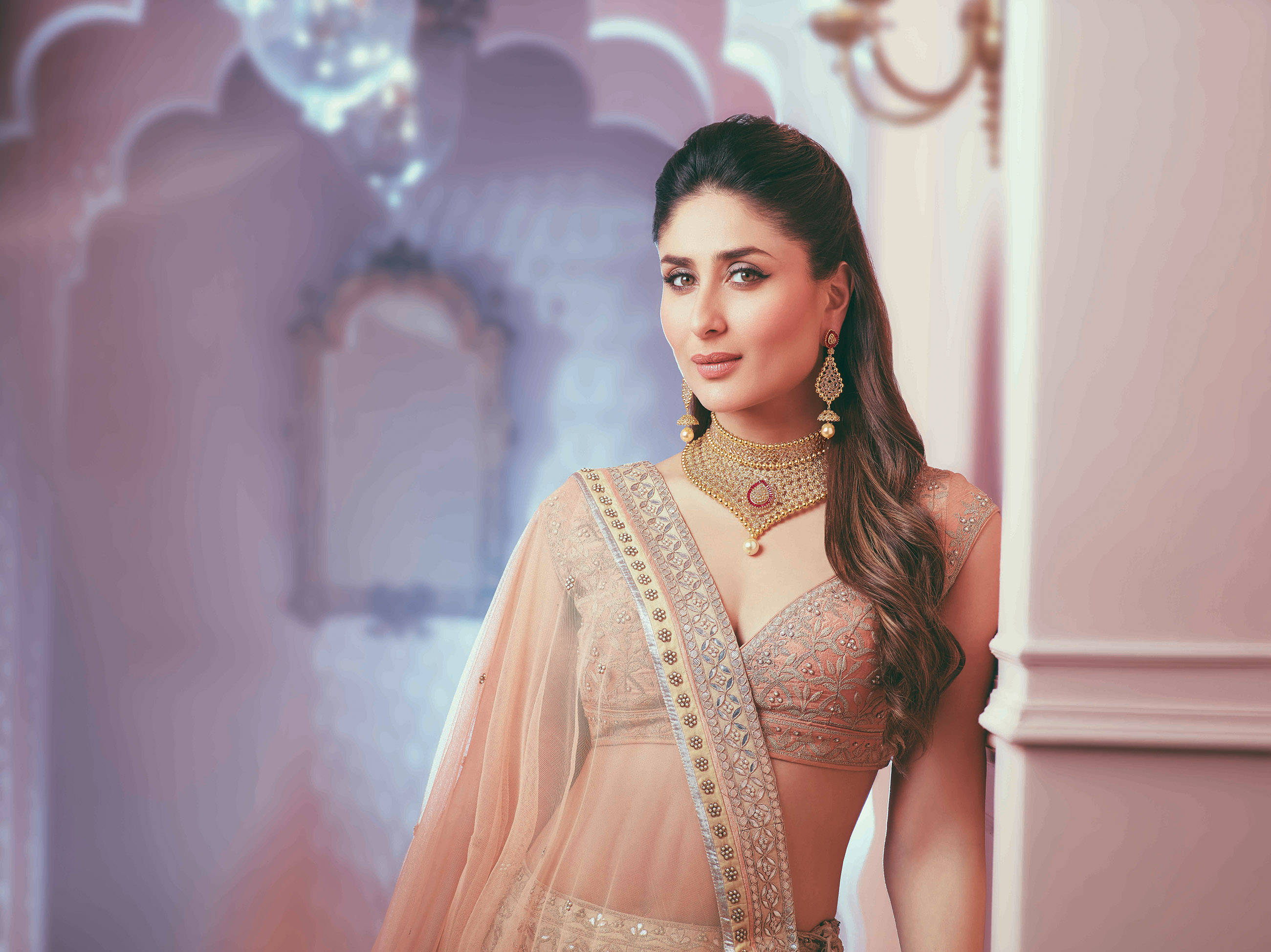 Kareena 桌布 壁紙 — 一位身穿華麗蜜桃色長裙和黃金首飾的女性，優雅地站在柔和燈光的粉紅色宮殿房間中。